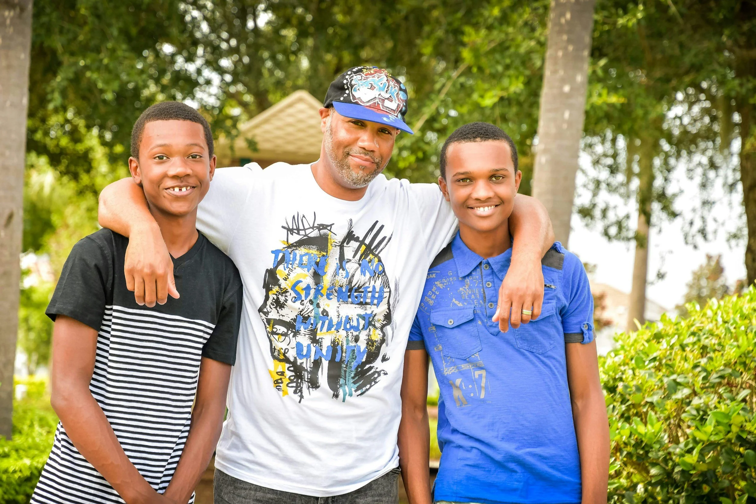 A man with two boys standing outdoors in a park with trees in the background. The man has gray facial hair and is wearing a white t-shirt with a graphic design, a black baseball cap with colorful designs, and has his arms around the boys. Both boys are smiling, one wearing a black and white striped t-shirt and the other in a blue shirt.