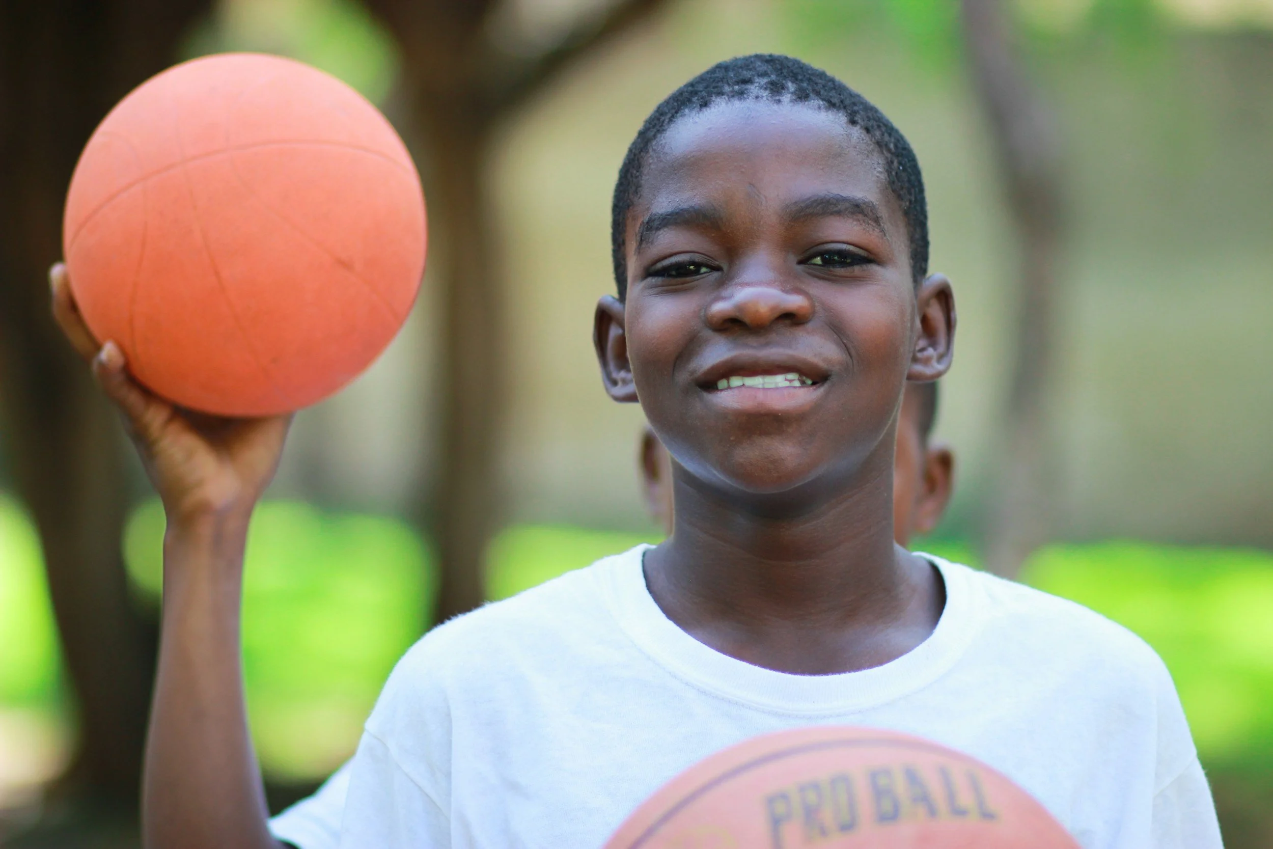 A young boy holding an orange basketball in his right hand, standing outdoors with trees and greenery in the background, wearing a white t-shirt, smiling slightly.