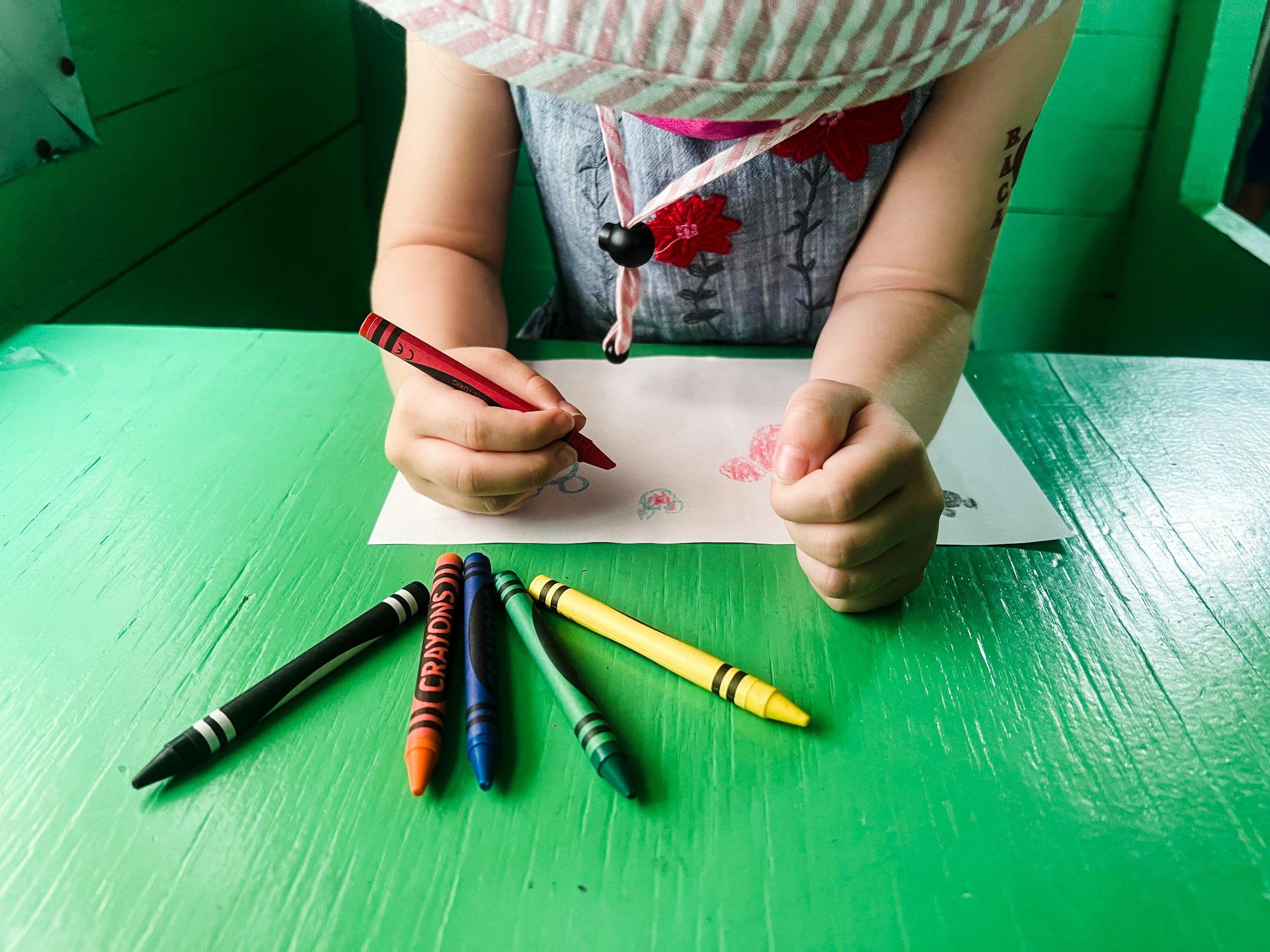 A young child wearing a striped shirt drawing with a red crayon on a piece of paper, with four additional colorful crayons on a bright green table.