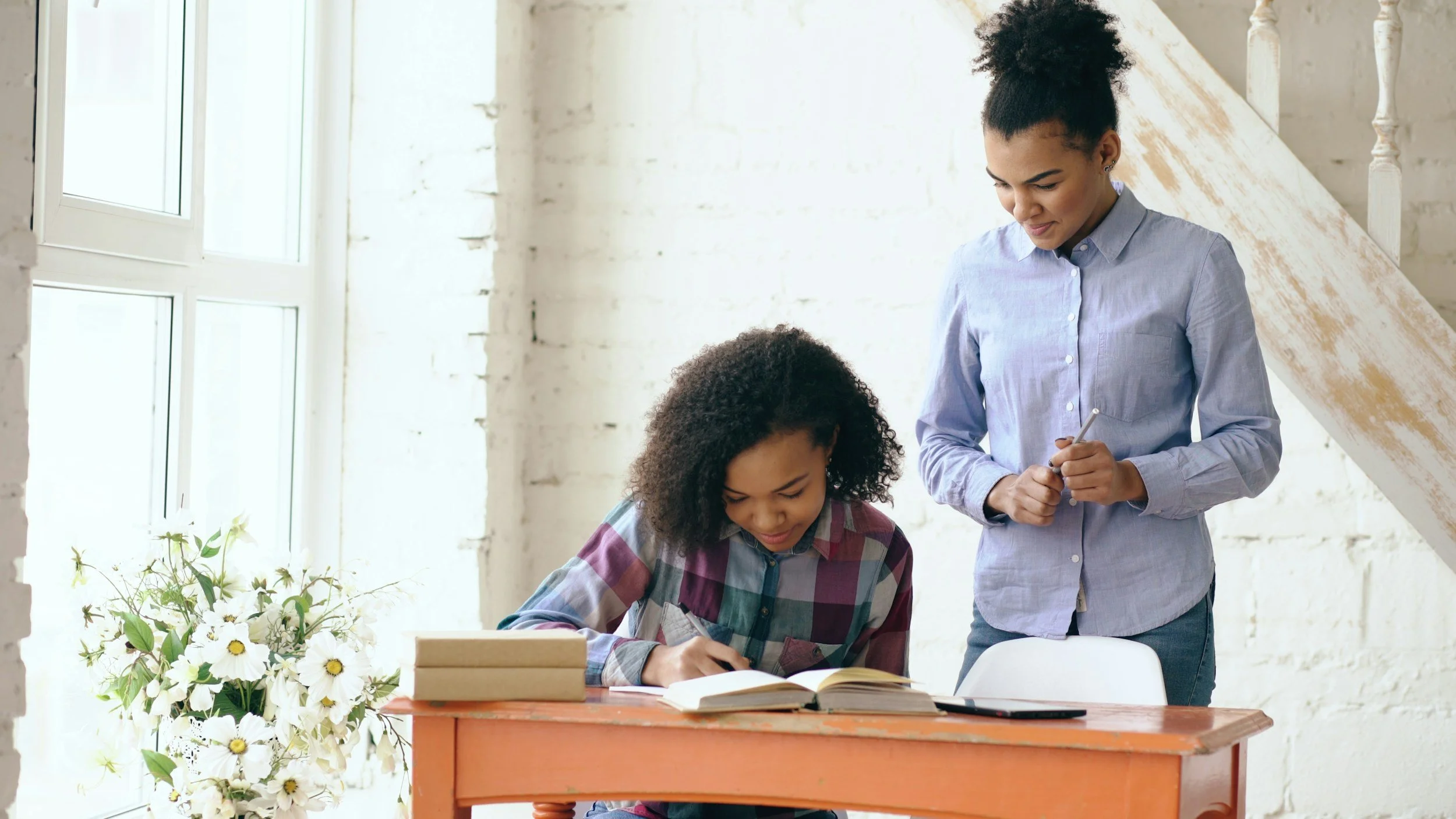 Two women working together at a desk with books, notebook, and pen in a bright room with large windows and white brick walls.
