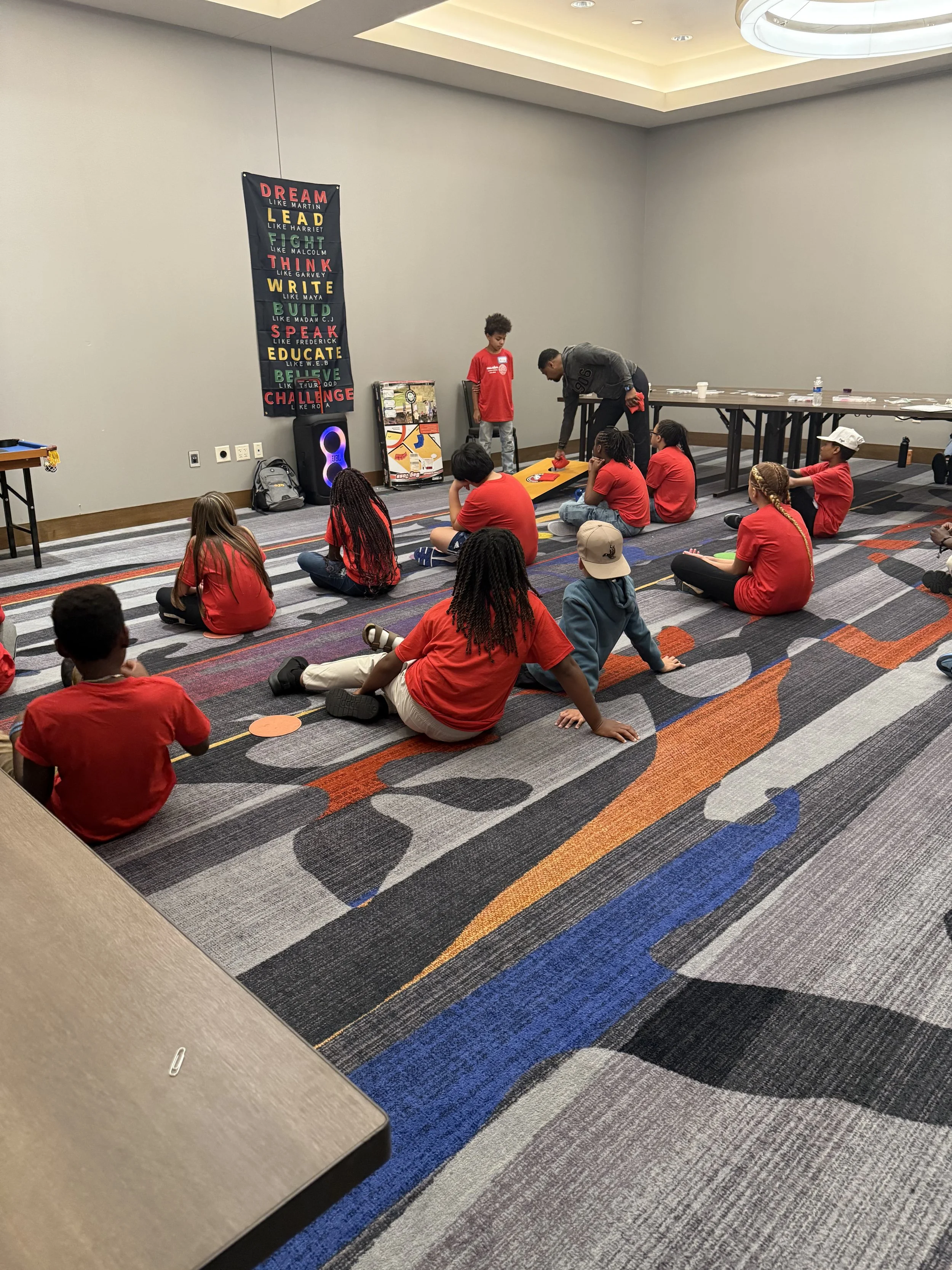 Children sitting on the carpet in a room, listening to a man who is demonstrating a game or activity at the front. The children are wearing red shirts, and some are sitting cross-legged while others are lying down. There is a colorful sign with motivational words on the wall and tables in the background.