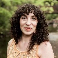 Portrait of a woman with curly dark hair, wearing a sleeveless yellow top, standing outdoors with greenery in the background.