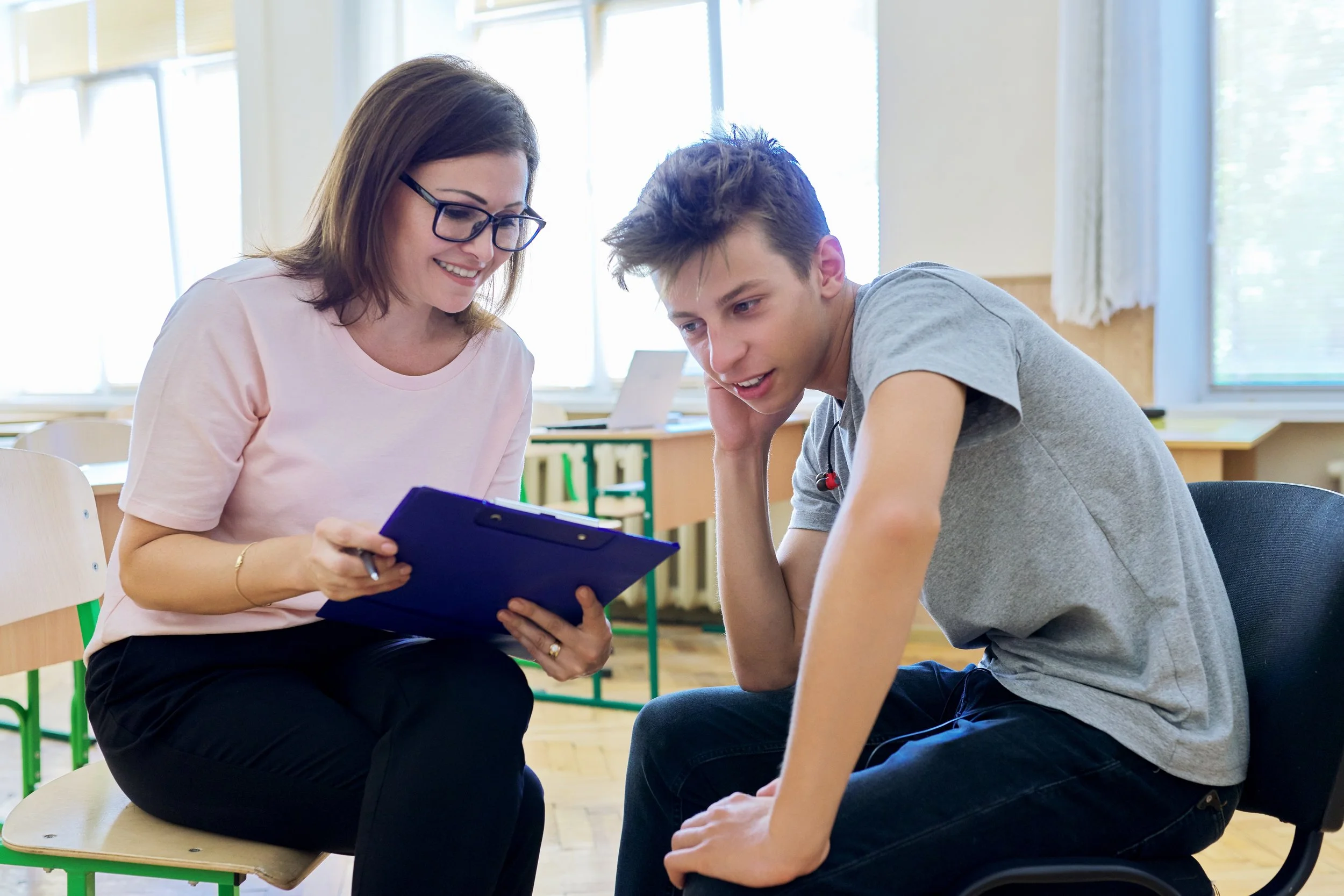 A woman with glasses and a pink shirt smiling while showing a notebook to a teenage boy with messy hair in a classroom.