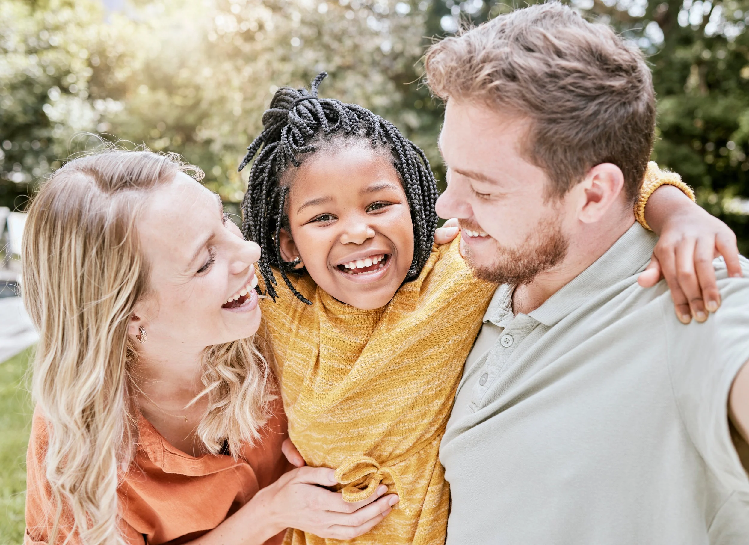 Family of three smiling and hugging outdoors, with a girl in the middle, two adults on either side, sunlight behind trees.