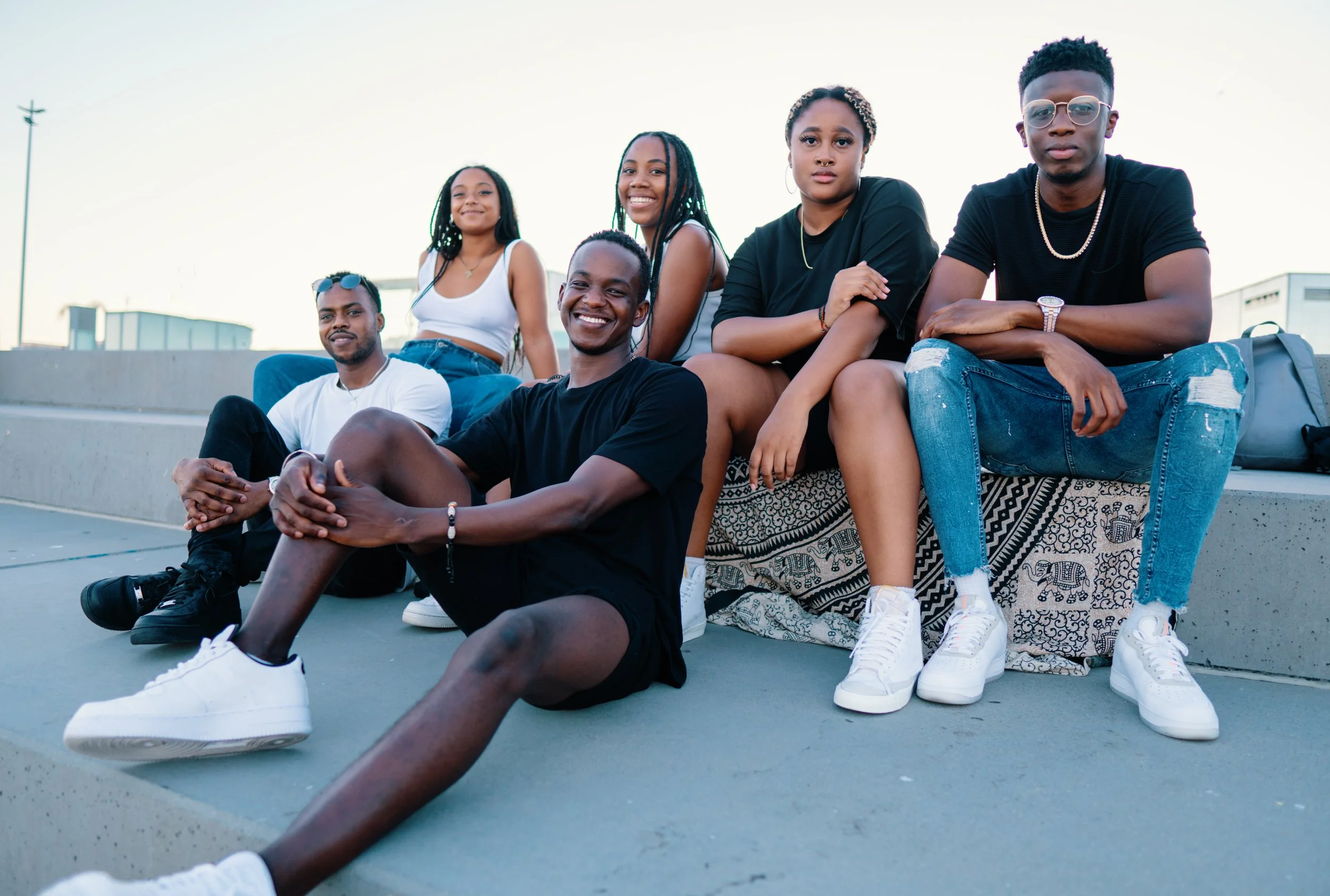 Group of six young adults sitting together on a concrete ledge outdoors, smiling and enjoying each other's company during sunset.