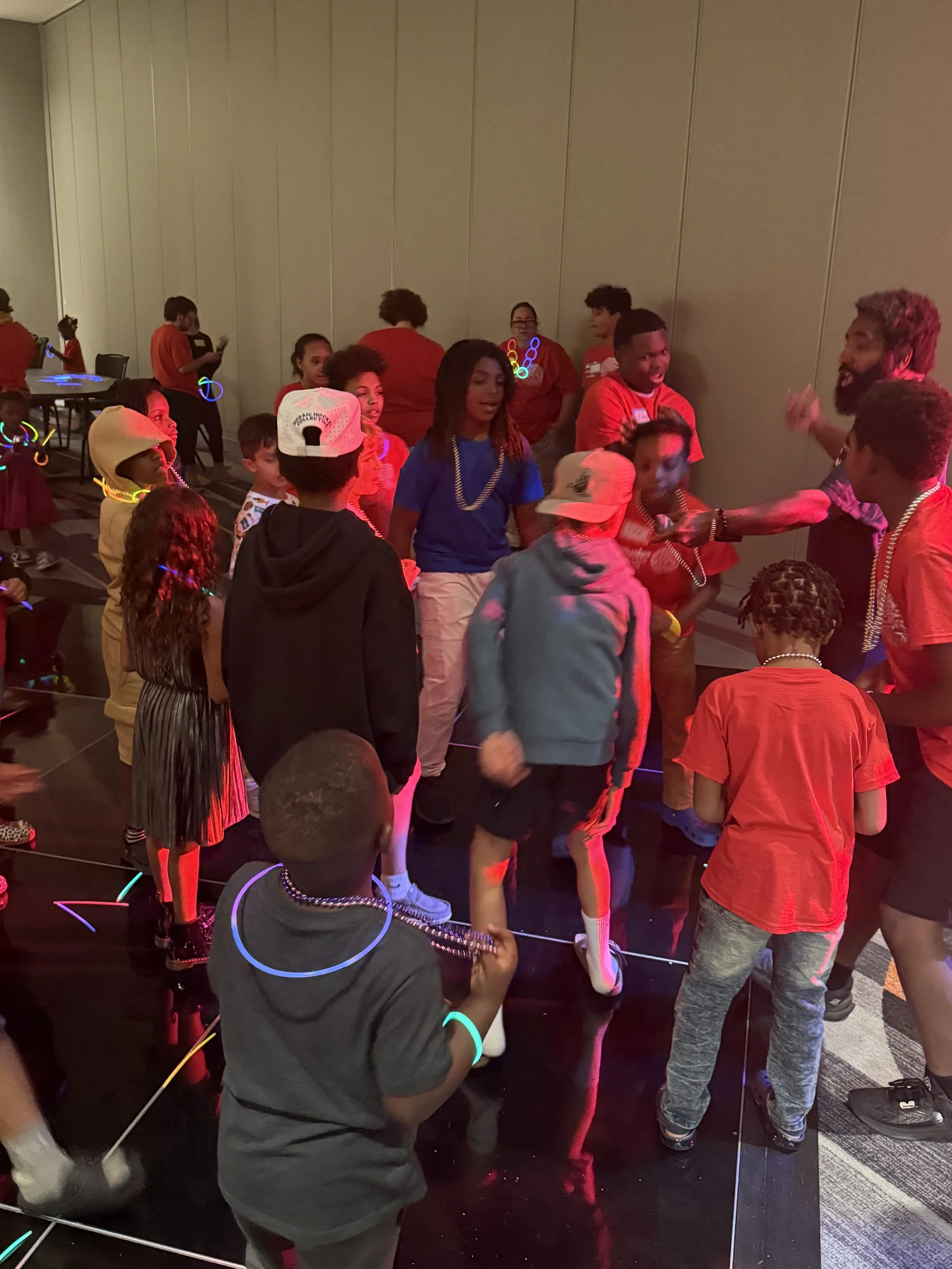 Children and adults dancing at a party with glow necklaces and bracelets in a dimly lit room.