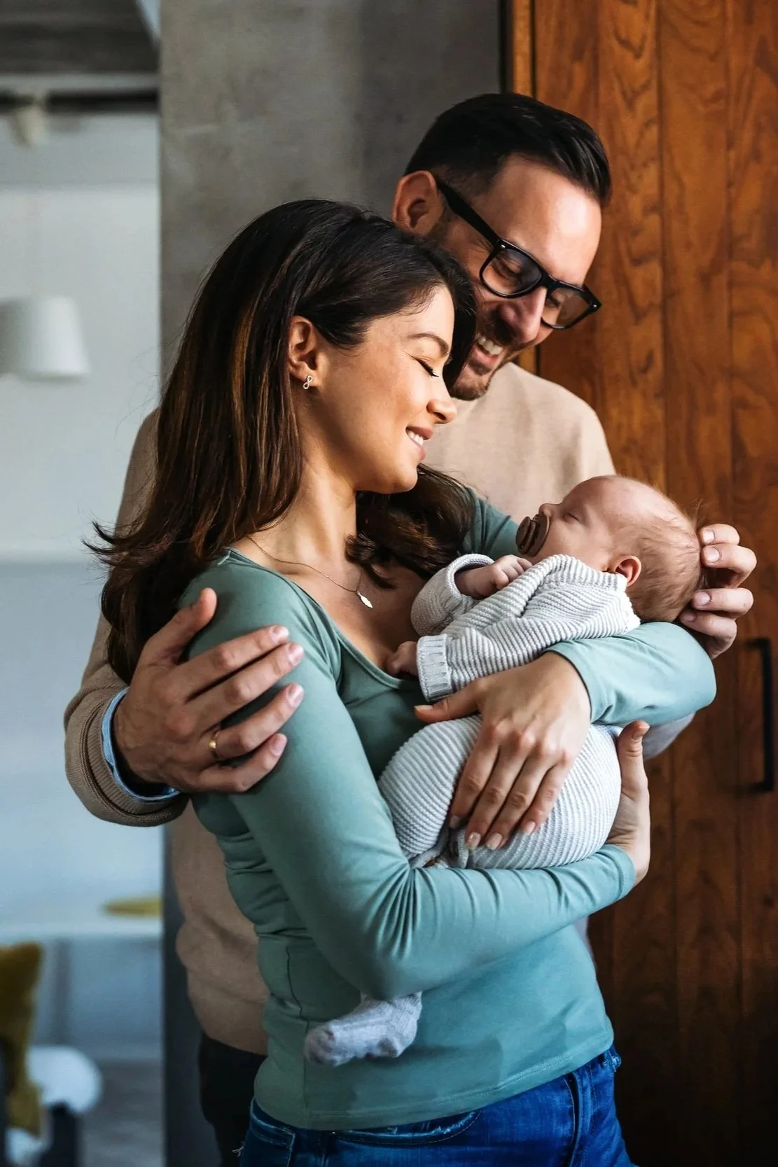 A smiling woman holding a baby with pacifier in mouth, with a man hugging her from behind, all sharing a joyful moment.