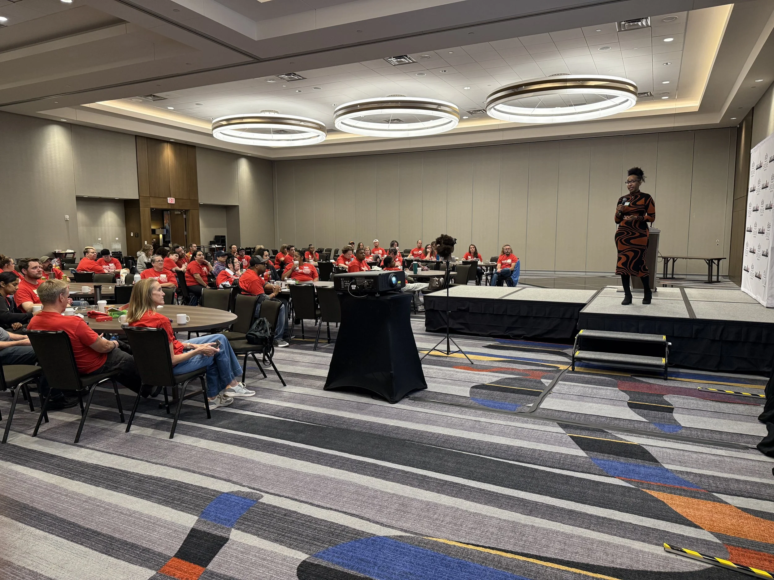 A woman speaking on stage to an audience in a conference room with many attendees dressed in red shirts.