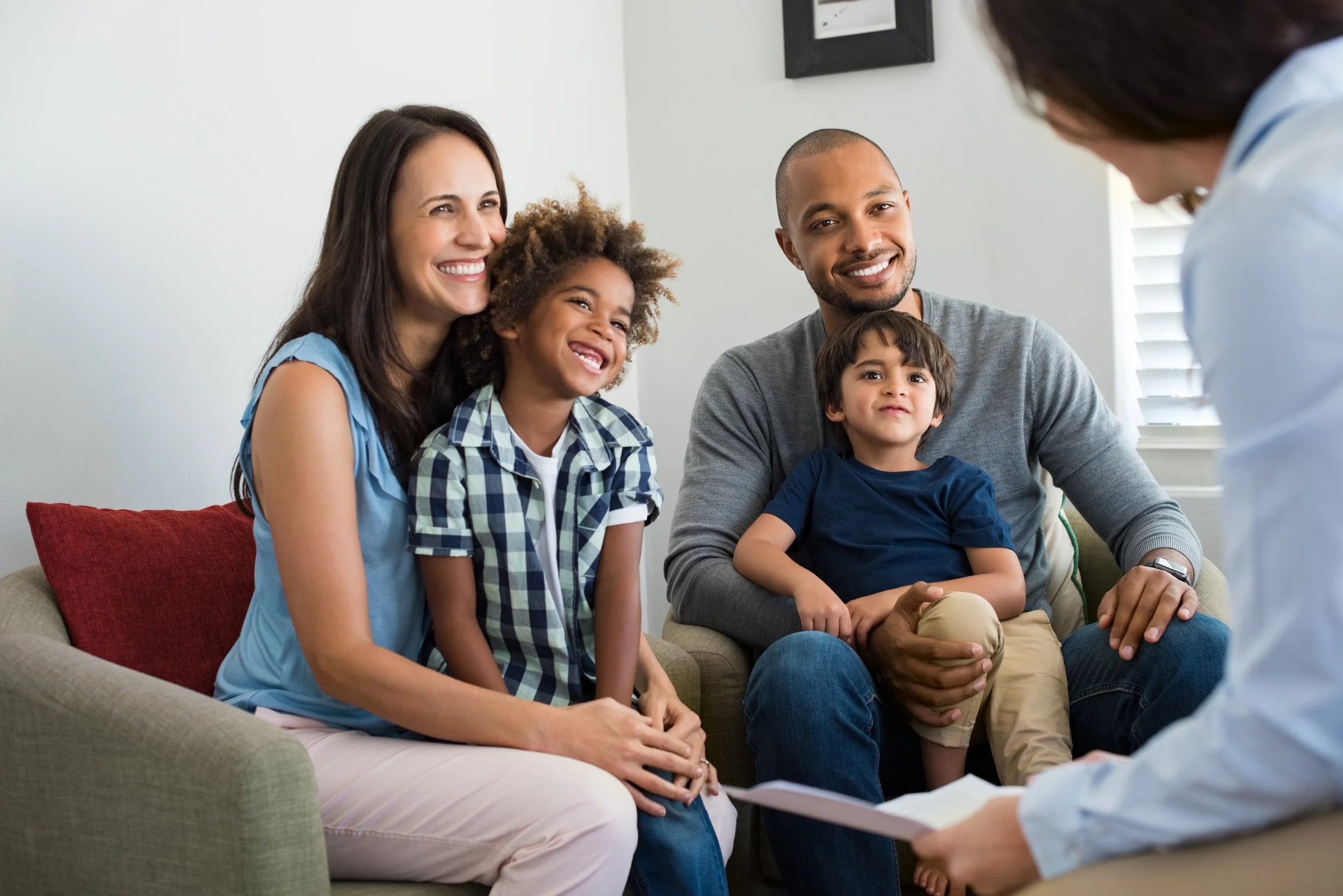 A family of four sitting on a couch during a medical appointment, talking to a doctor who is holding a clipboard. The family appears happy and engaged.
