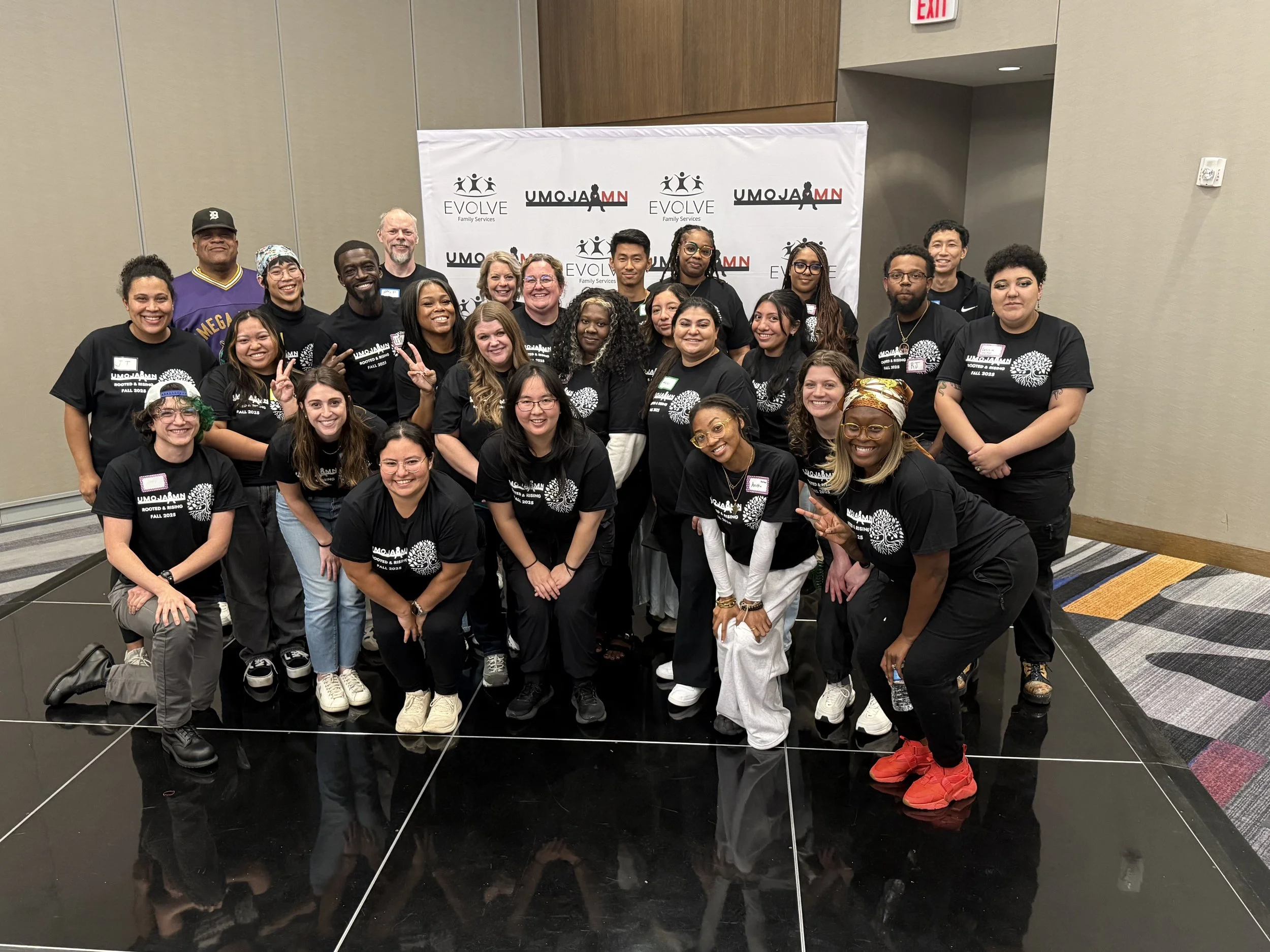 Group of diverse people posing together on a stage in front of a backdrop with logos. They are wearing black T-shirts with white text and designs, smiling and making various hand gestures.