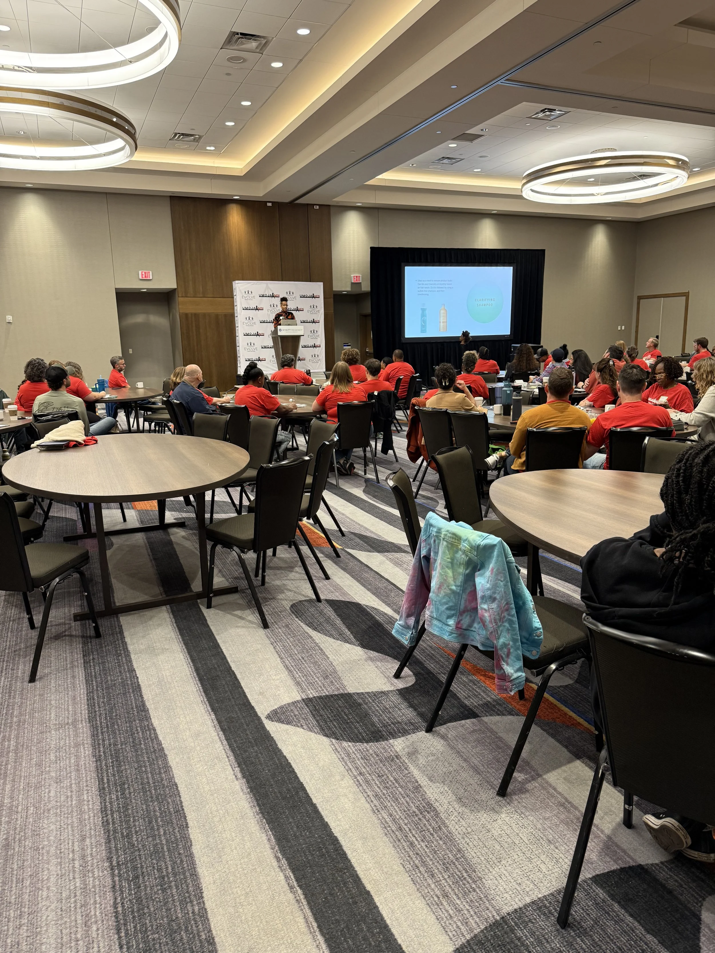 A conference room with many attendees sitting at round tables, some wearing red shirts, listening to a speaker at a presentation. A large screen displays a slide with spray bottles. The room features modern lighting and a patterned carpet.