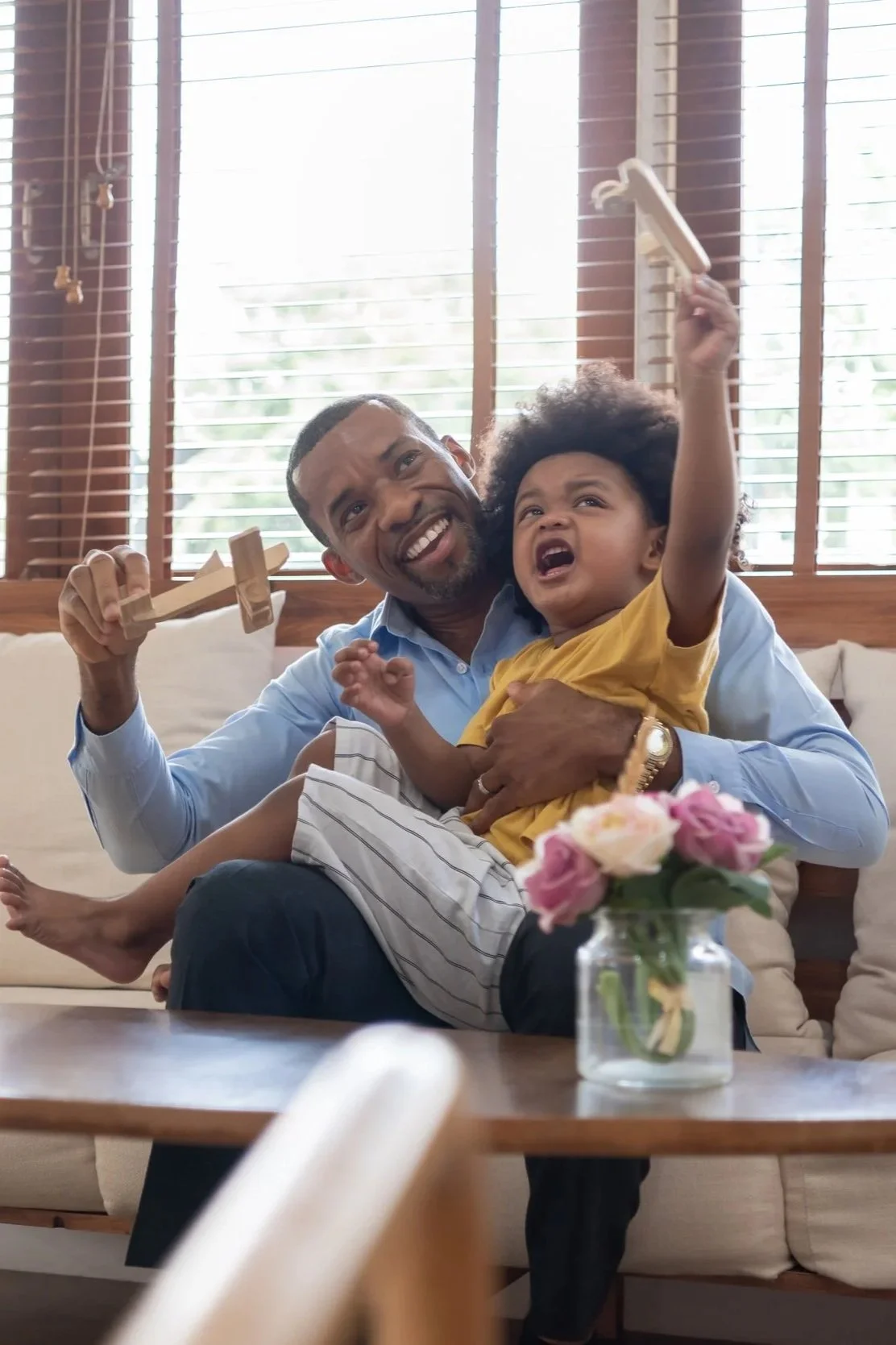 Man playing with a young girl on a beige couch in a bright living room with wooden blinds, a vase of pink roses on a wooden table in the foreground.