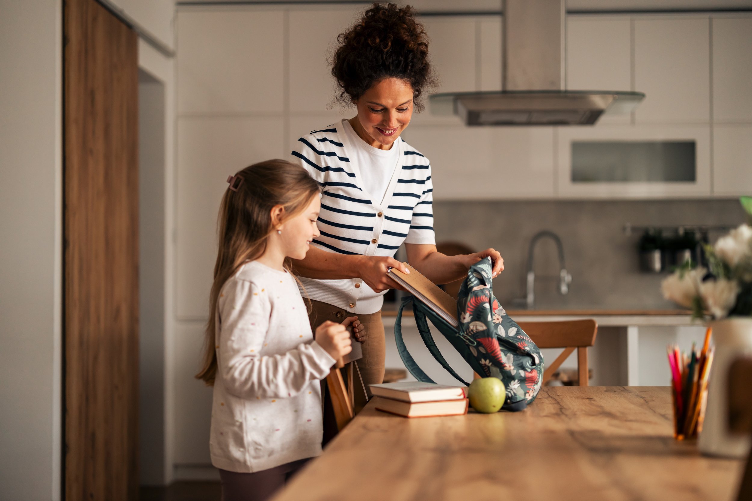 A woman and a girl pack a backpack in a kitchen