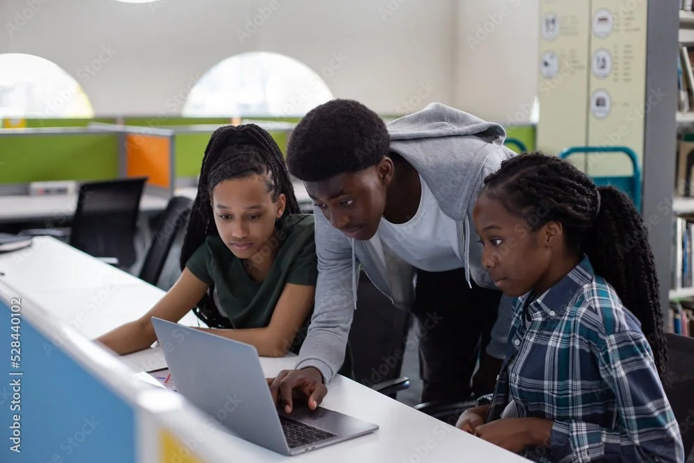 Three children in a library looking at a laptop screen, two girls and a boy leaning over to view the laptop