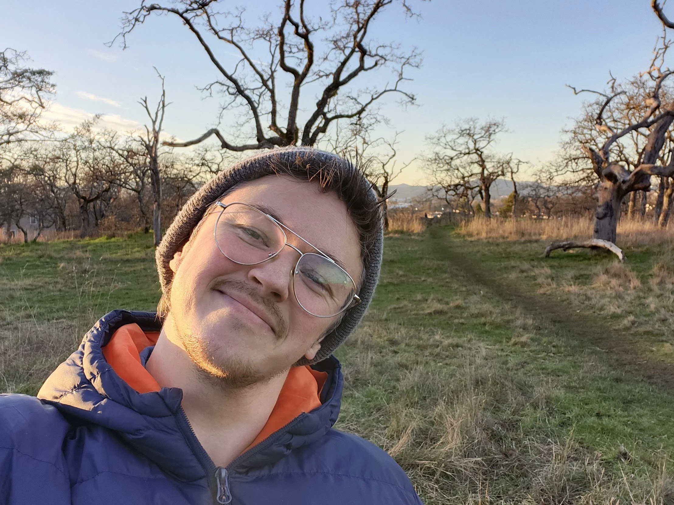 a white trans-masculine person standing in a garry oak meadow in winter. They are wearing glasses and have a beard