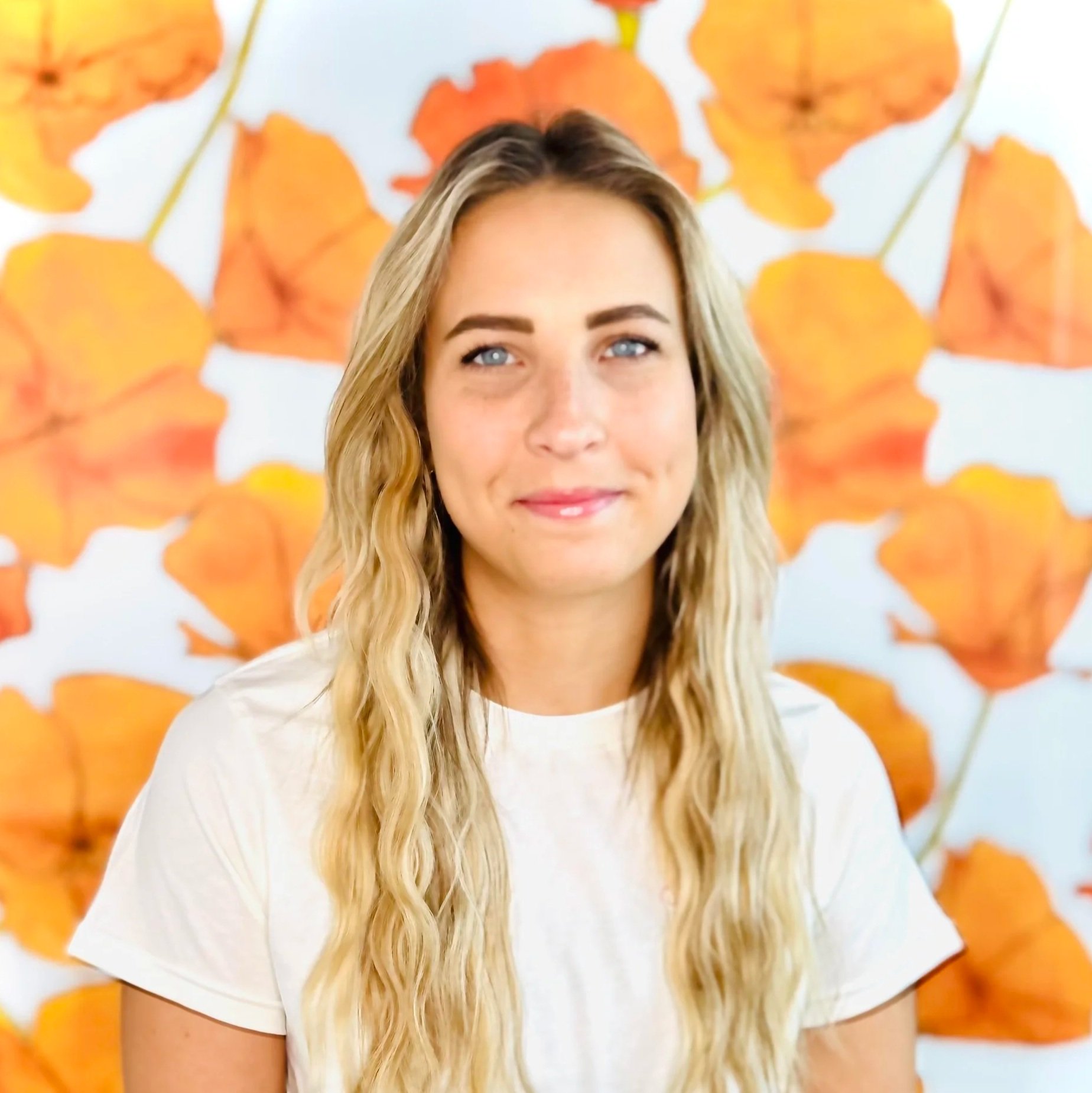 A young woman with long, wavy blonde hair and blue eyes smiling in front of a backdrop decorated with orange leaves.