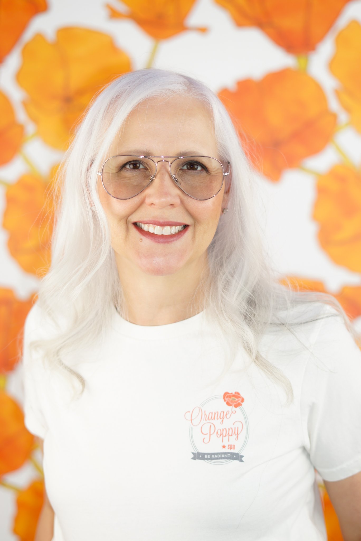 A Licensed Esthetician with long white hair wearing sunglasses and a white t-shirt with an 'Orange Poppy Spa' logo. She is smiling and standing in front of a background decorated with orange poppies.