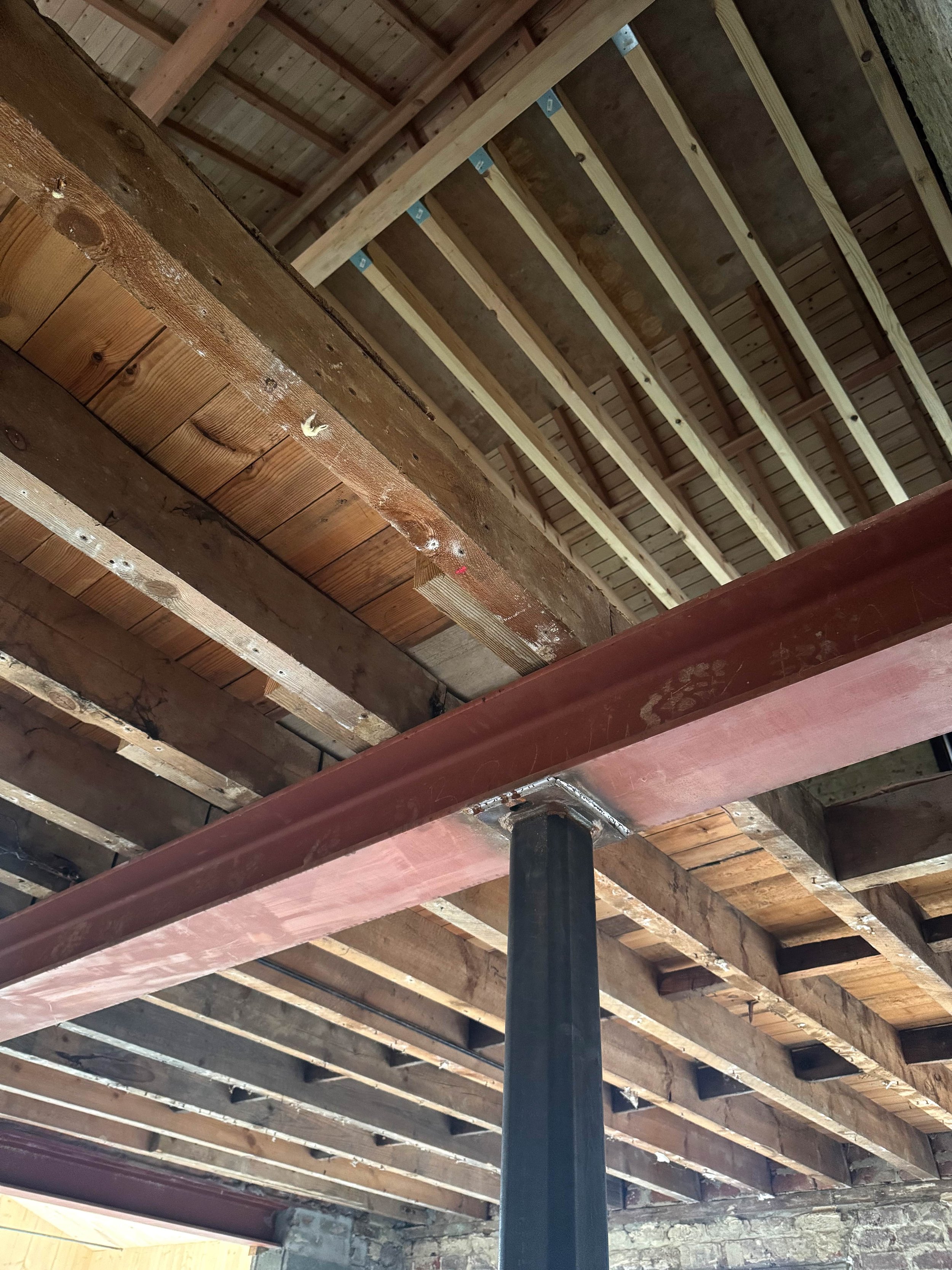 Interior view of a building under construction showing exposed wooden ceiling beams, metal support columns, and wooden framing for the floor above.