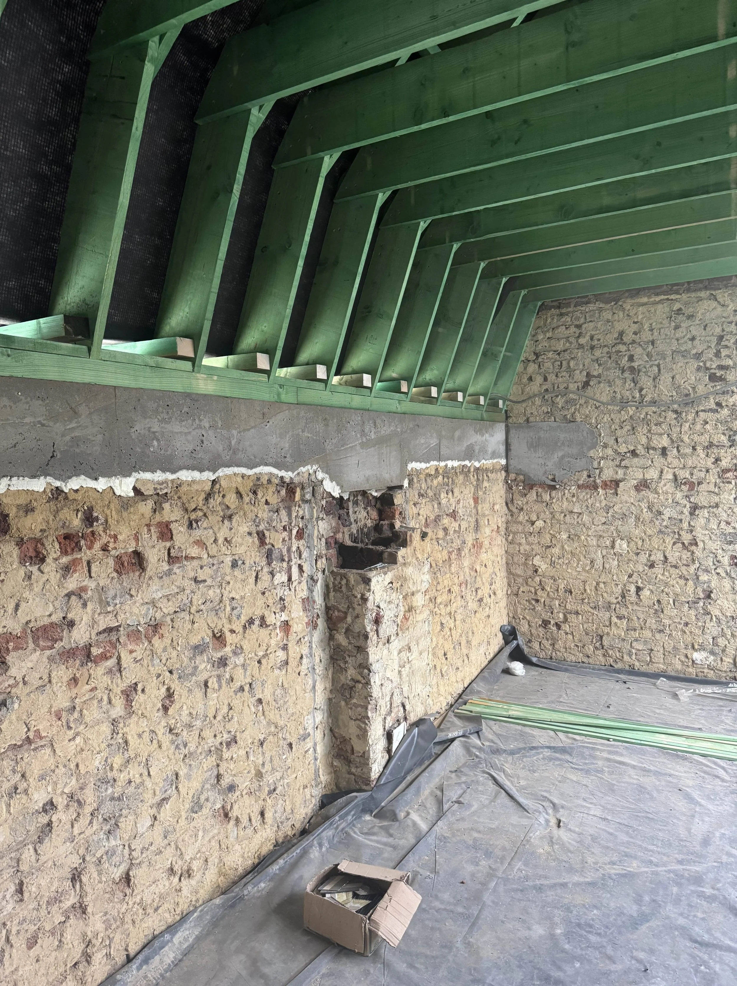 Interior of a building under renovation showing exposed brick and concrete wall with green wooden roof framing, construction materials on the floor, and plastic sheeting.