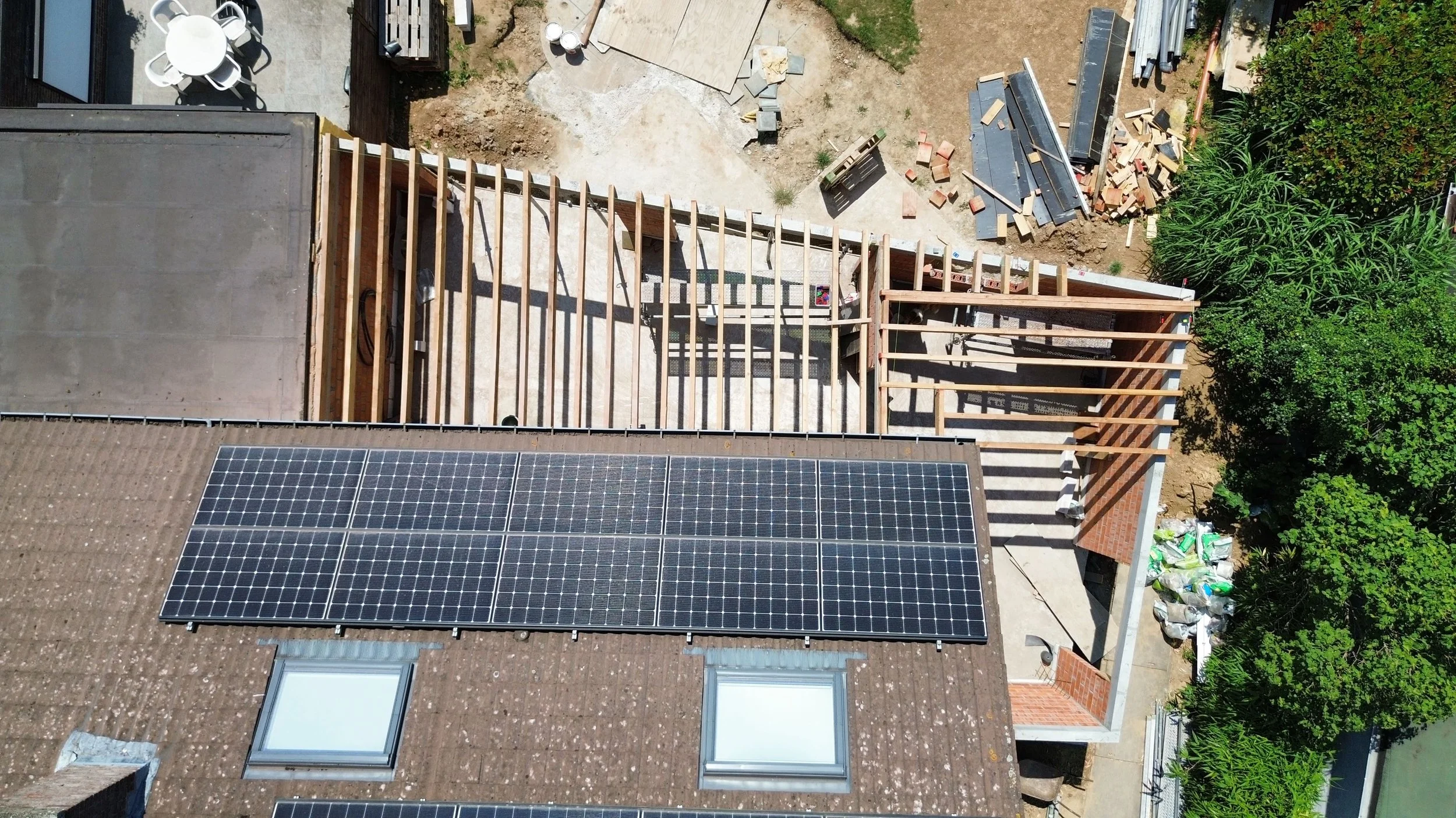 Aerial view of a house under construction with solar panels on the roof, and a backyard with a wooden deck, construction materials, and greenery.