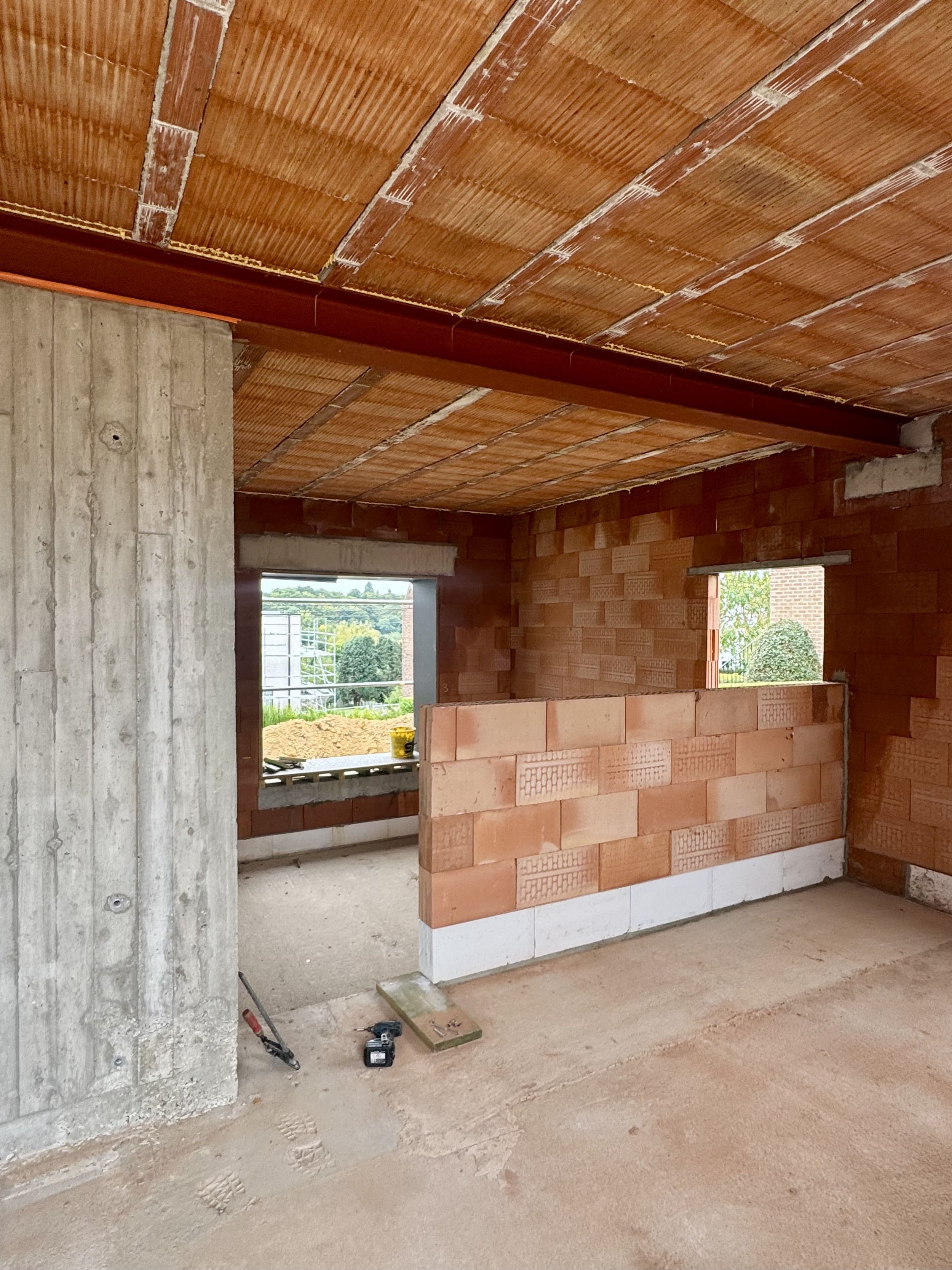 Interior view of a building under construction with unfinished brick walls, a large window opening, and construction tools on the floor.