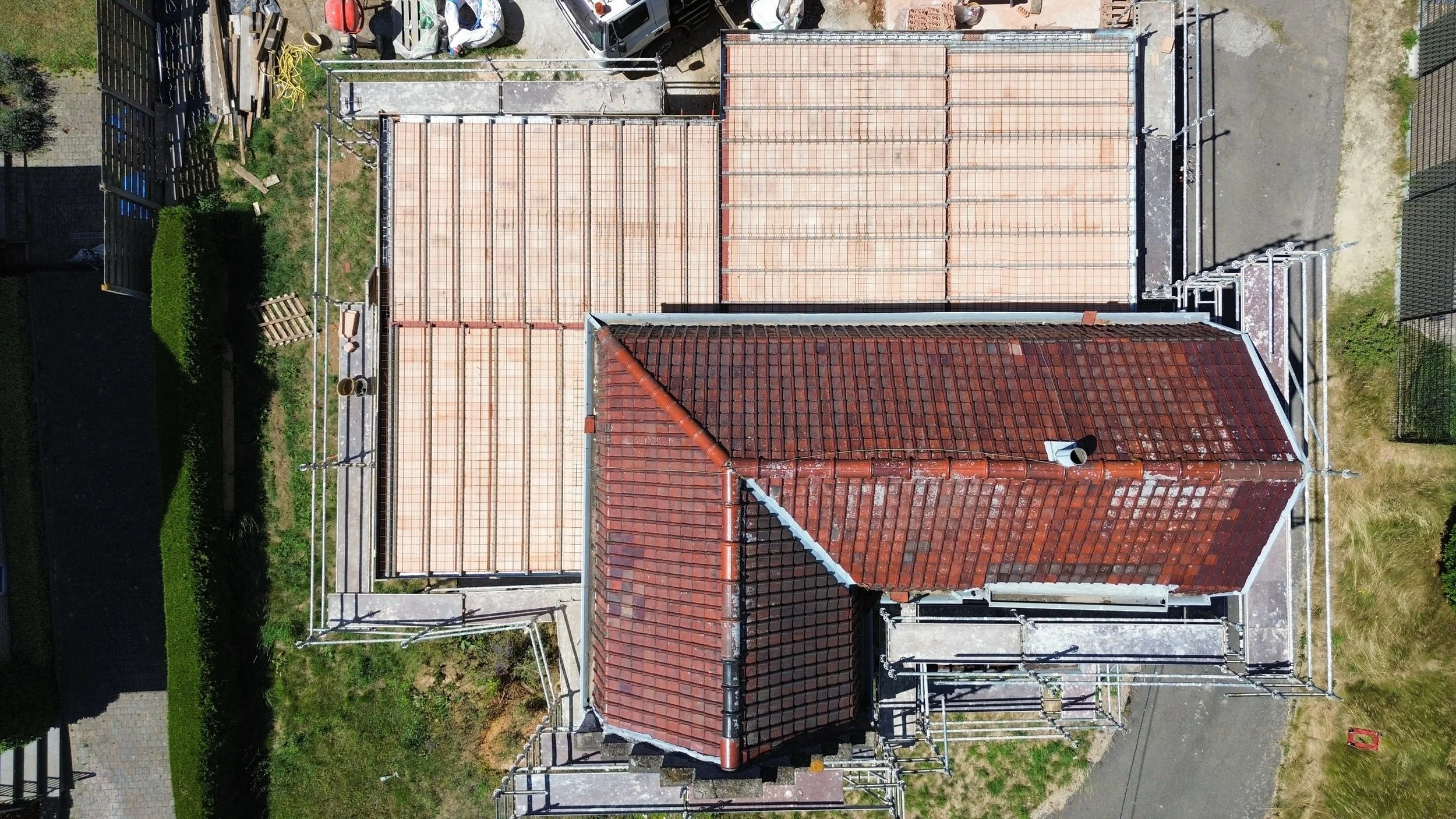 Top-down aerial view of a building under construction with a red tiled roof and scaffolding around it, adjacent to parking spaces and pathways.