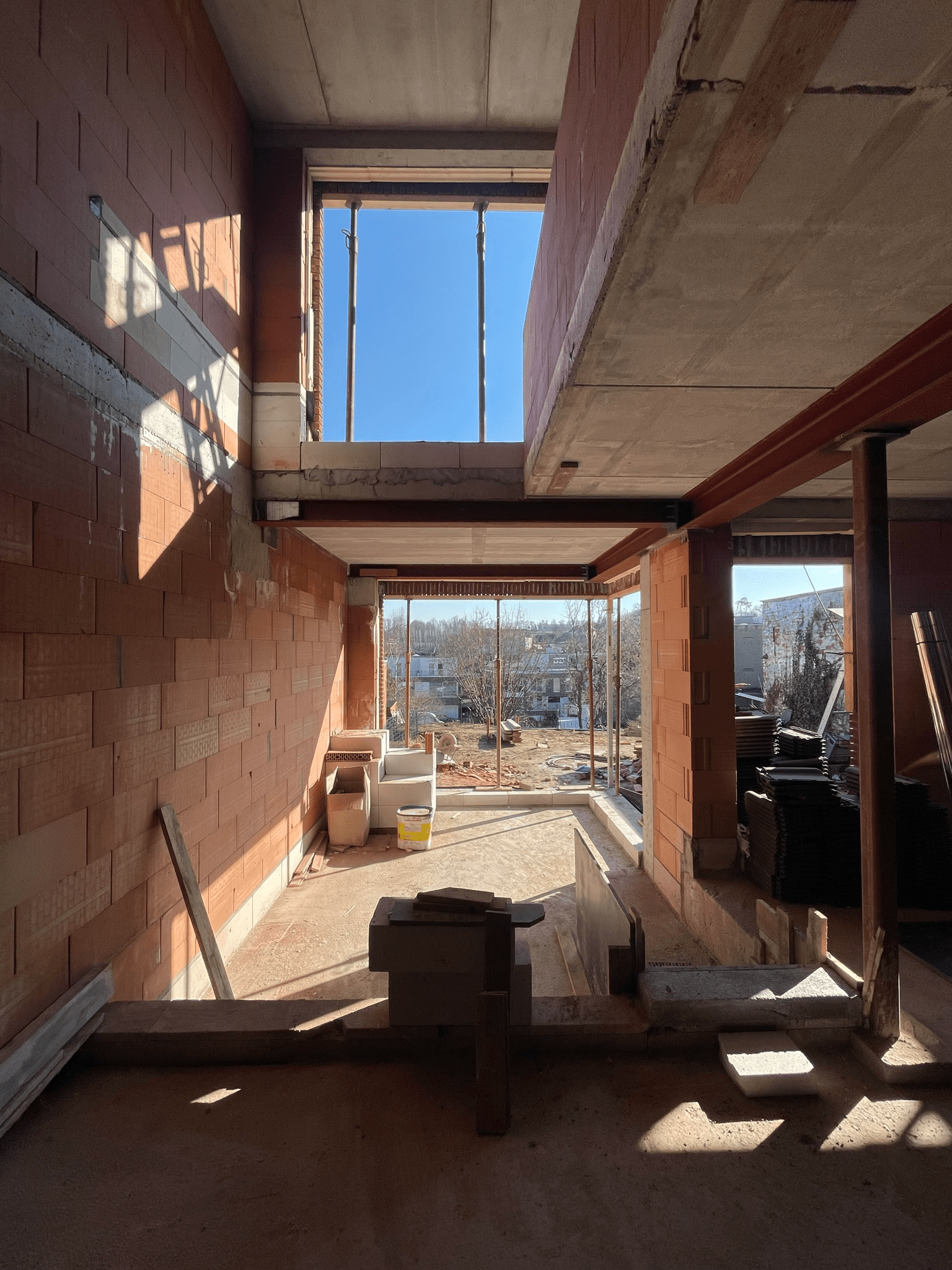 View of the interior of a building under construction with brick walls, concrete floors, and a large window opening showing a clear blue sky and an outdoor scene.