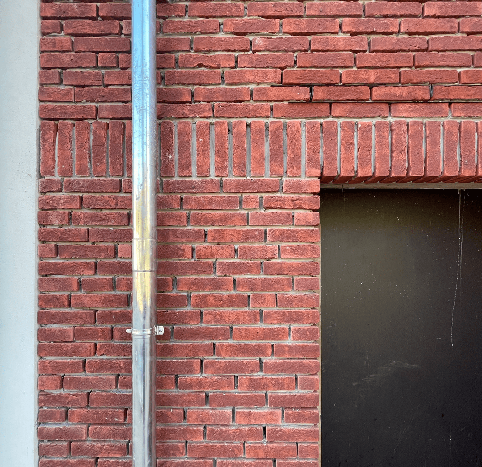 Close-up of a red brick wall with a metallic downspout pipe and a black opening on the right side.
