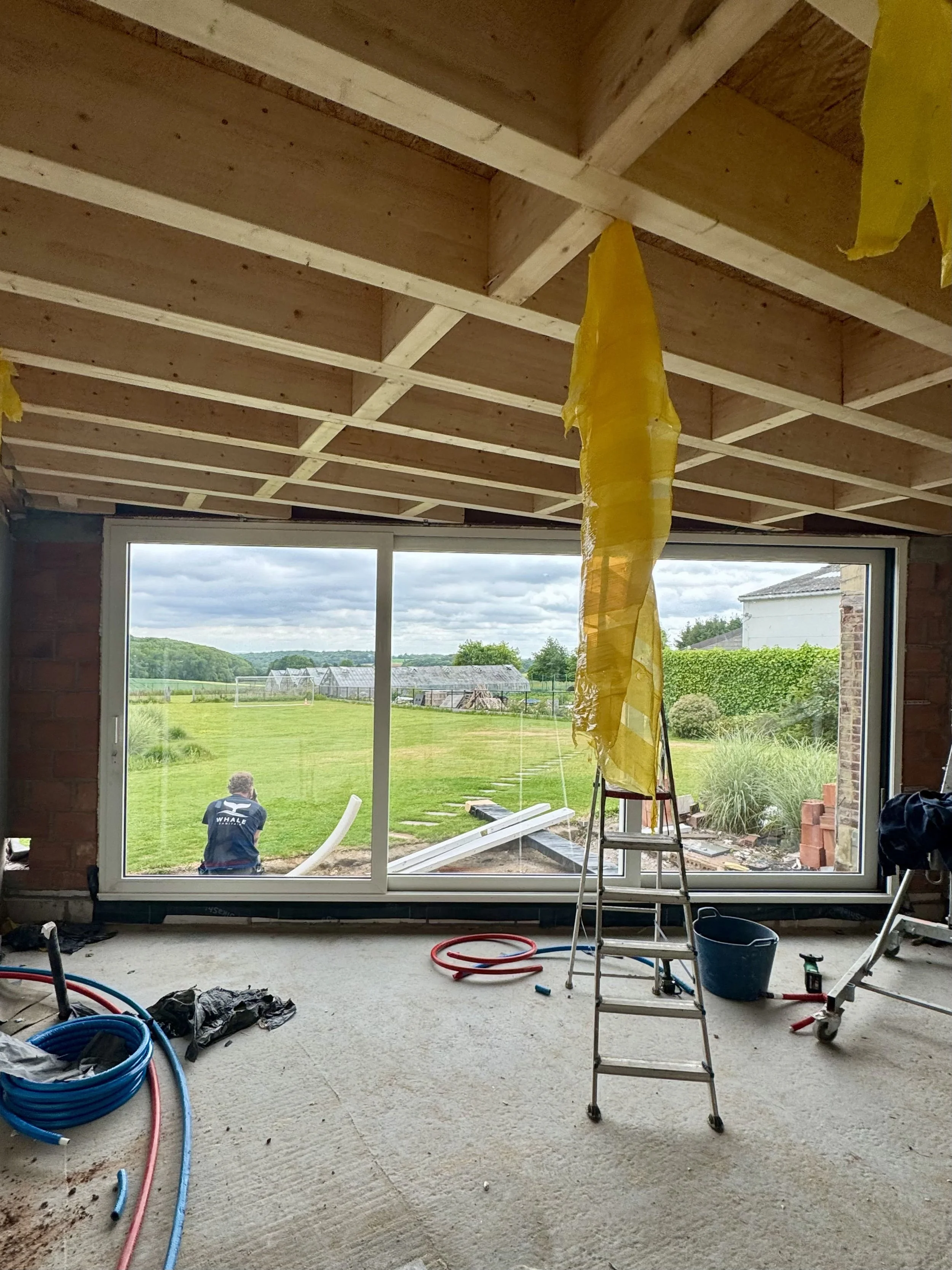 A construction site showing an unfinished room with a large window, a wood beam ceiling, a yellow plastic sheet hanging from the ceiling, and construction tools and materials scattered on the floor.
