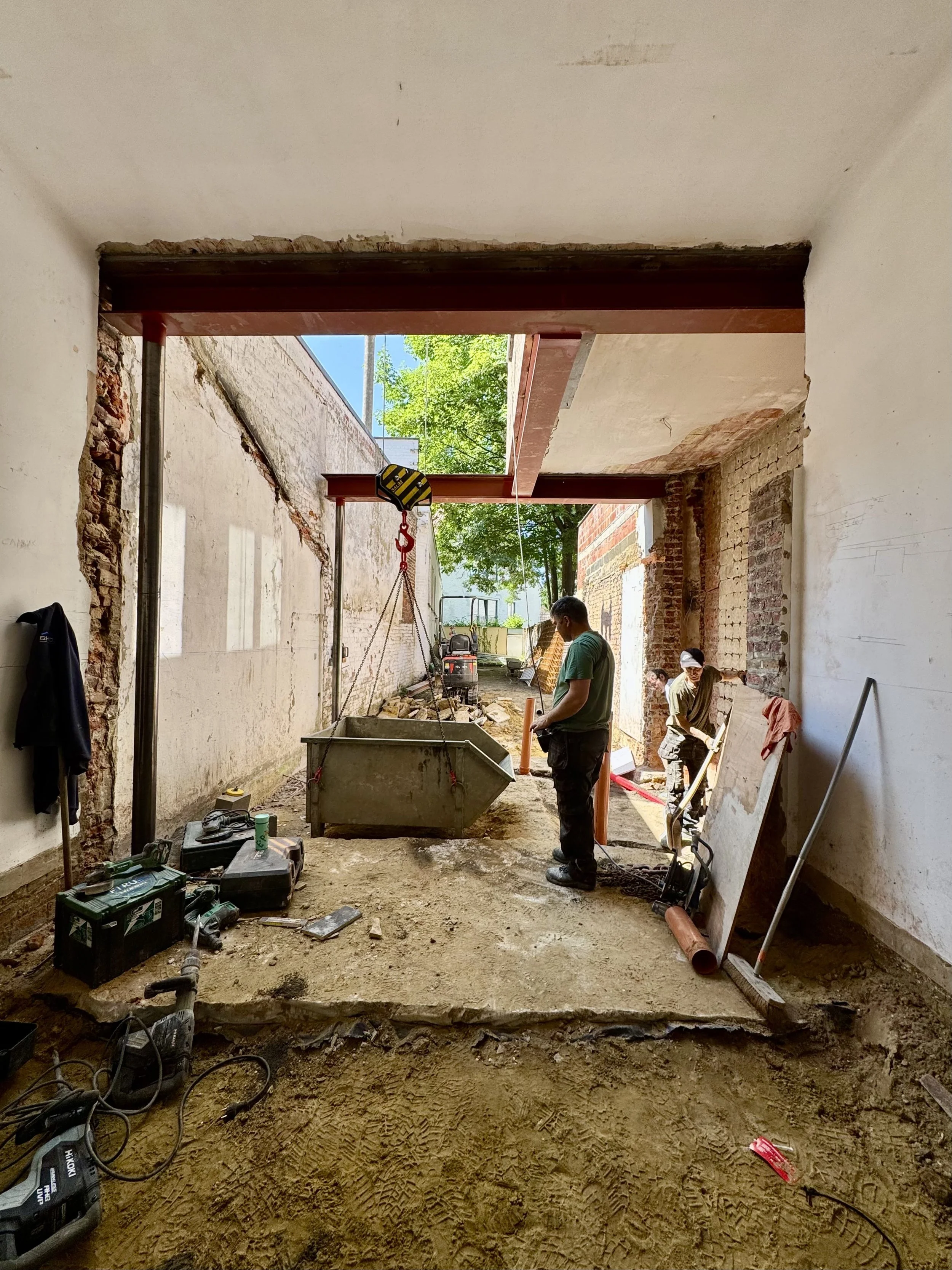 Construction workers renovate a building, installing foundation and support beams, with tools and equipment on the ground, and debris and bricks on the walls.