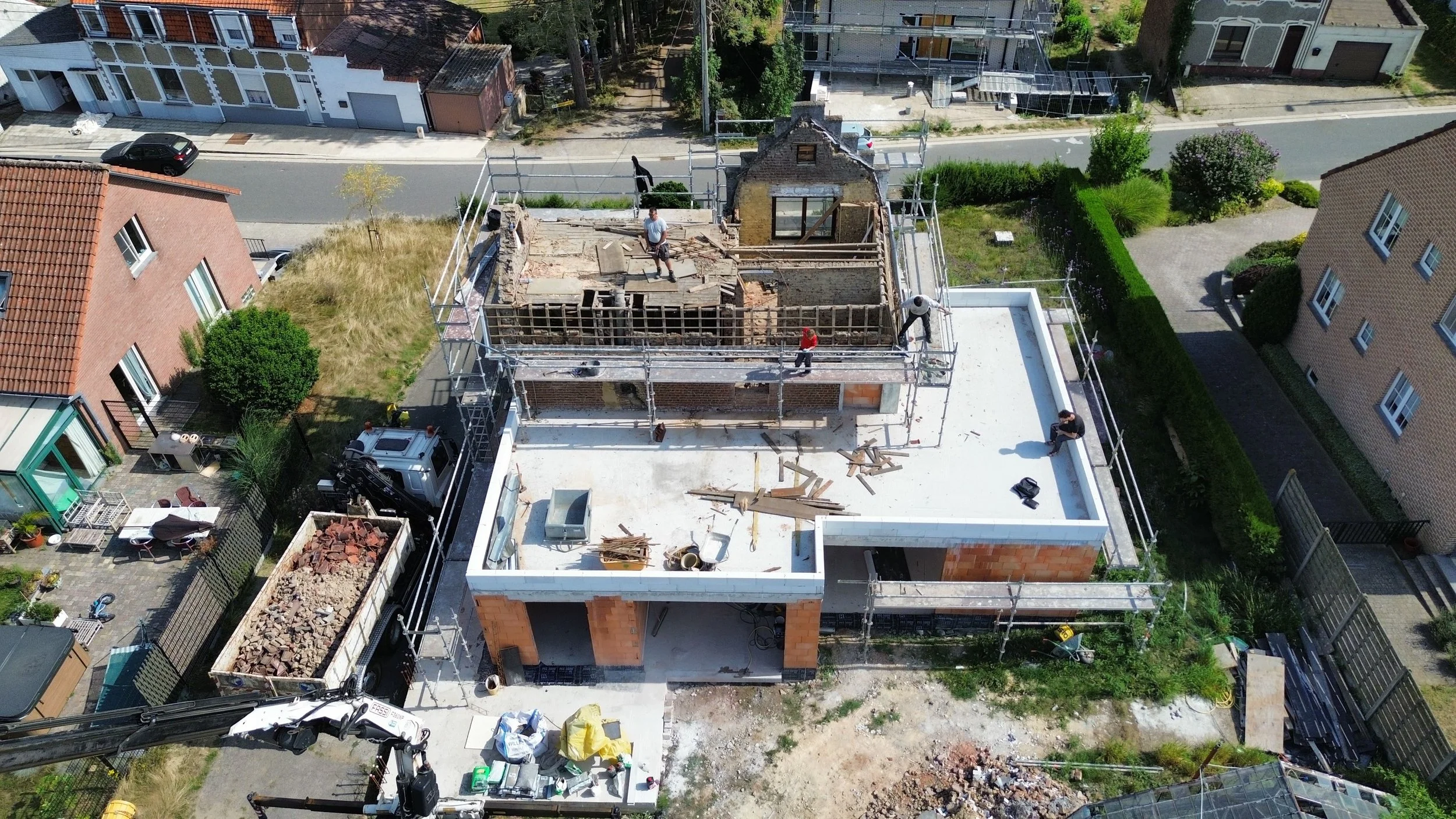 Aerial view of a house under construction with scaffolding, workers, building materials, and nearby parked cars, in a residential neighborhood with surrounding houses and greenery.