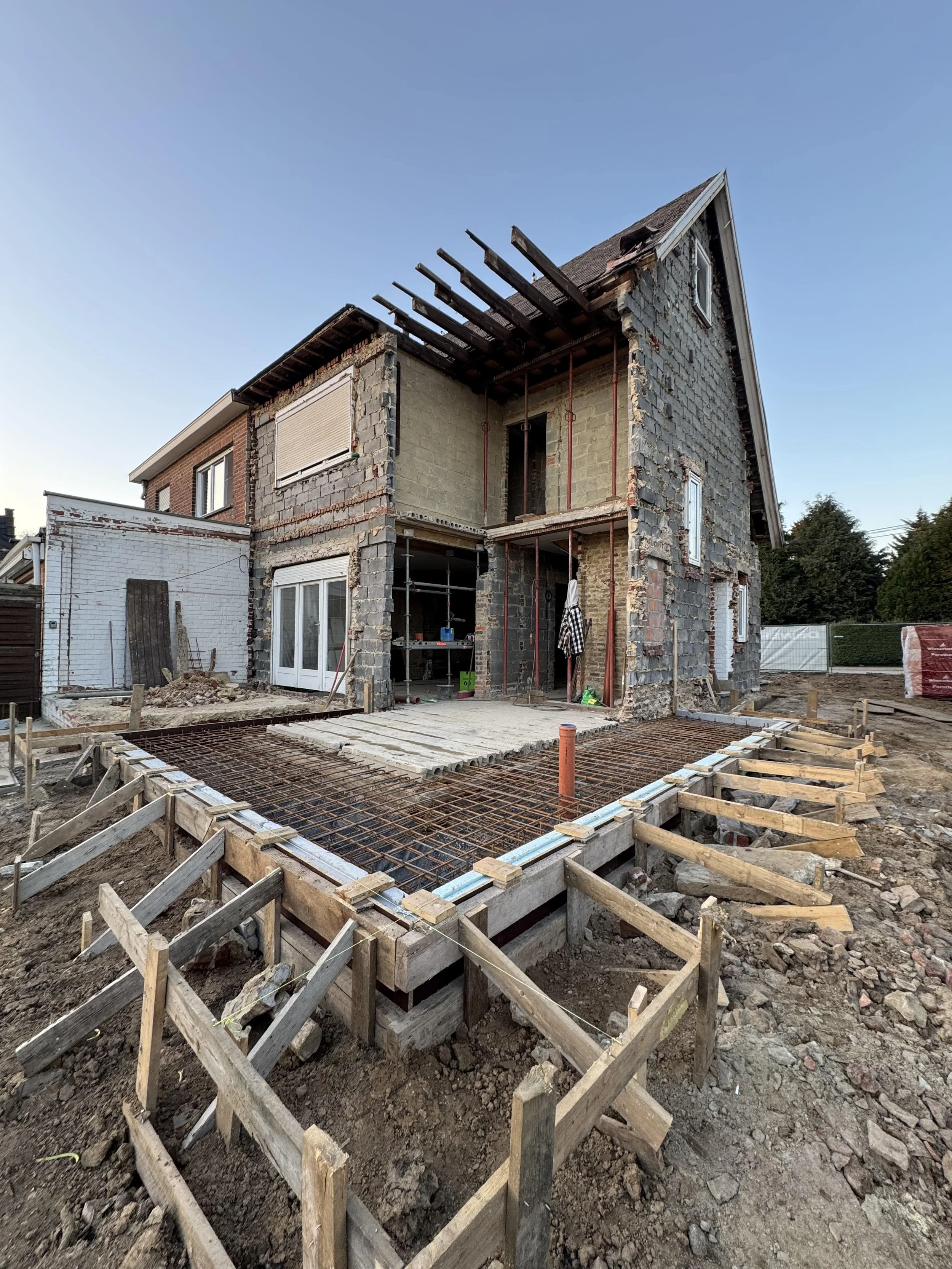 House under construction with exposed brick and stone walls, scaffolding, and a wooden platform in front, with construction materials around, on a clear day.