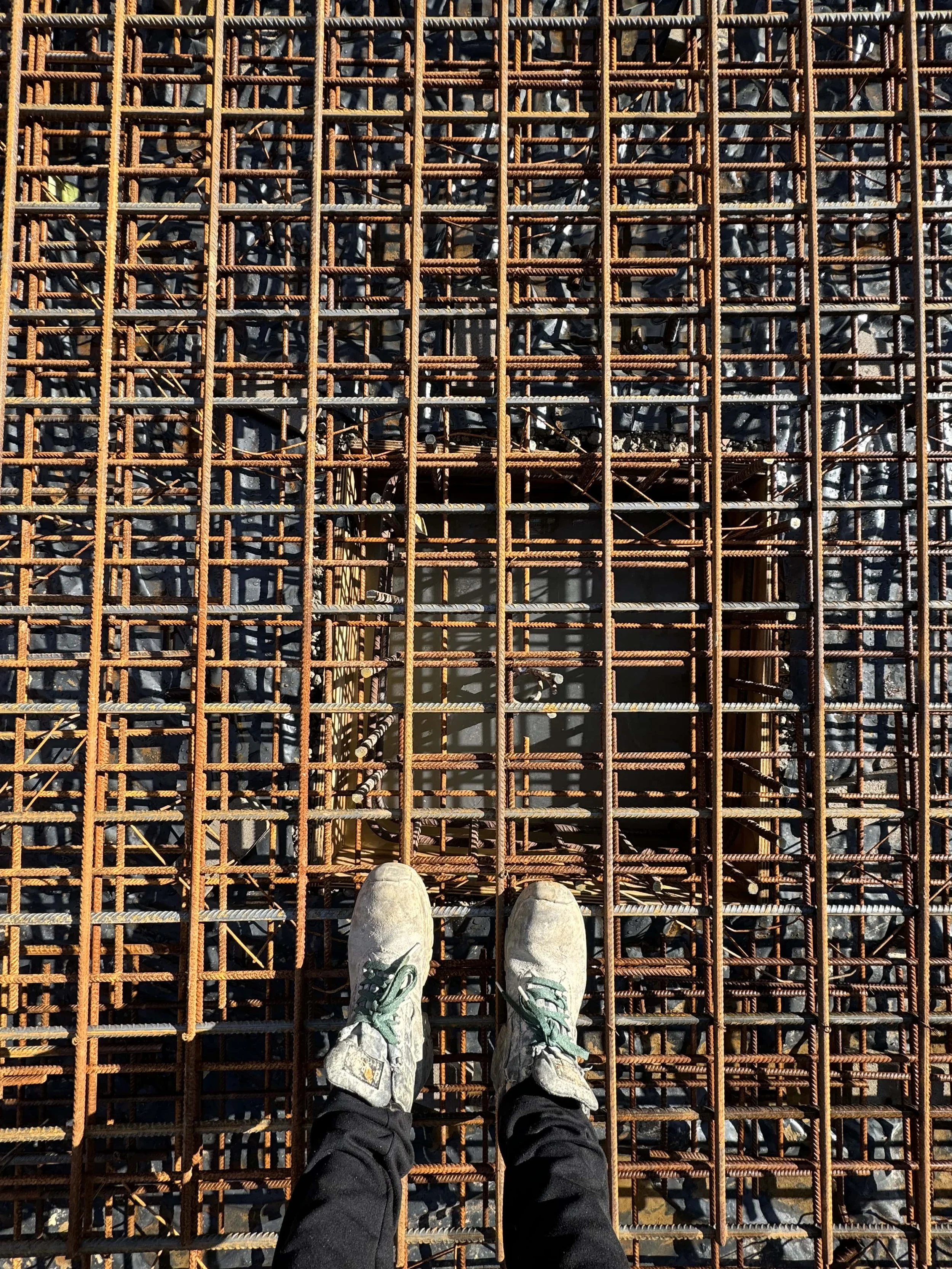A person wearing white sneakers and black pants standing on a rusted metal rebar grid at a construction site.