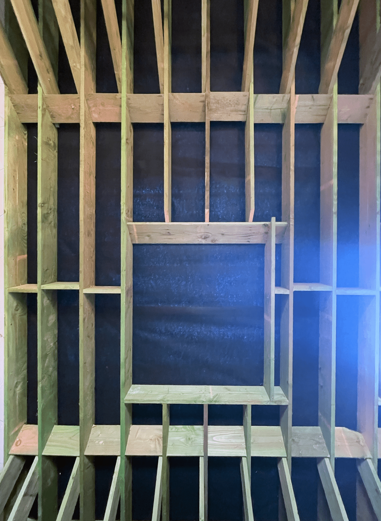 Interior view of a wooden framing structure with studs and a partial built-in frame or opening, possibly for a wall or window, inside a room under construction.
