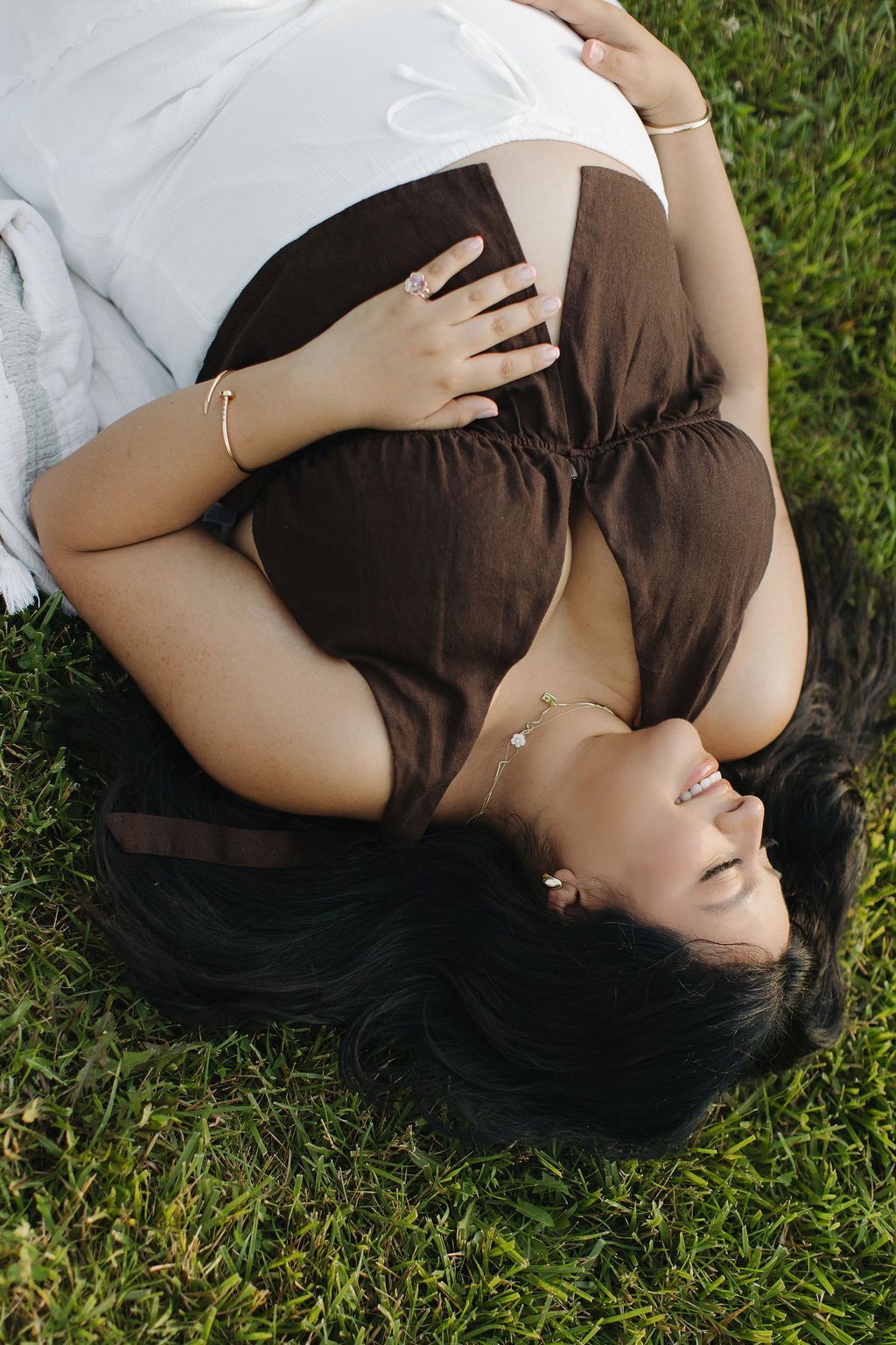 A woman lying on grass, smiling, with black hair, wearing a brown and white outfit, jewelry including a ring, bracelet, and necklace.