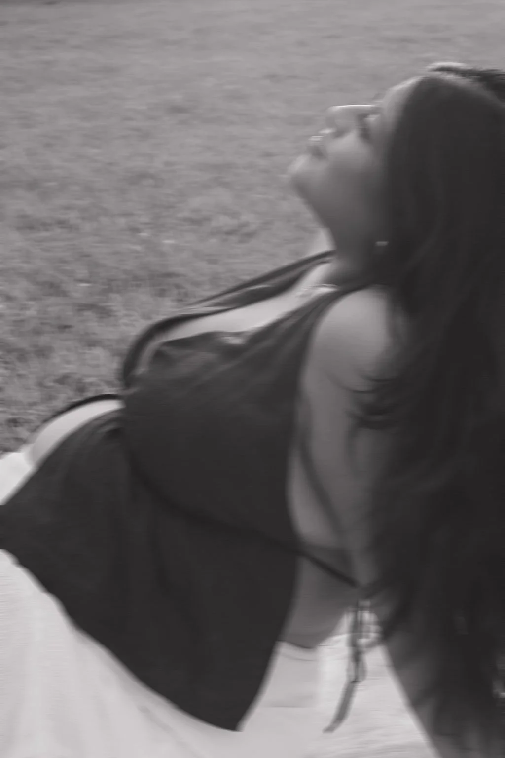 A woman with long dark hair lying on grass with her head tilted back and smiling.