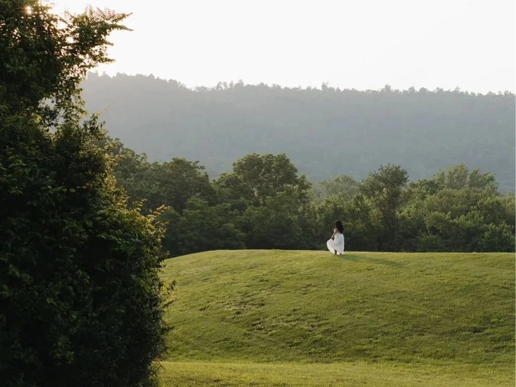 A maternity shoot of a woman in a white dress sits on a grassy hill overlooking a lush, green forest with mountains in the background.