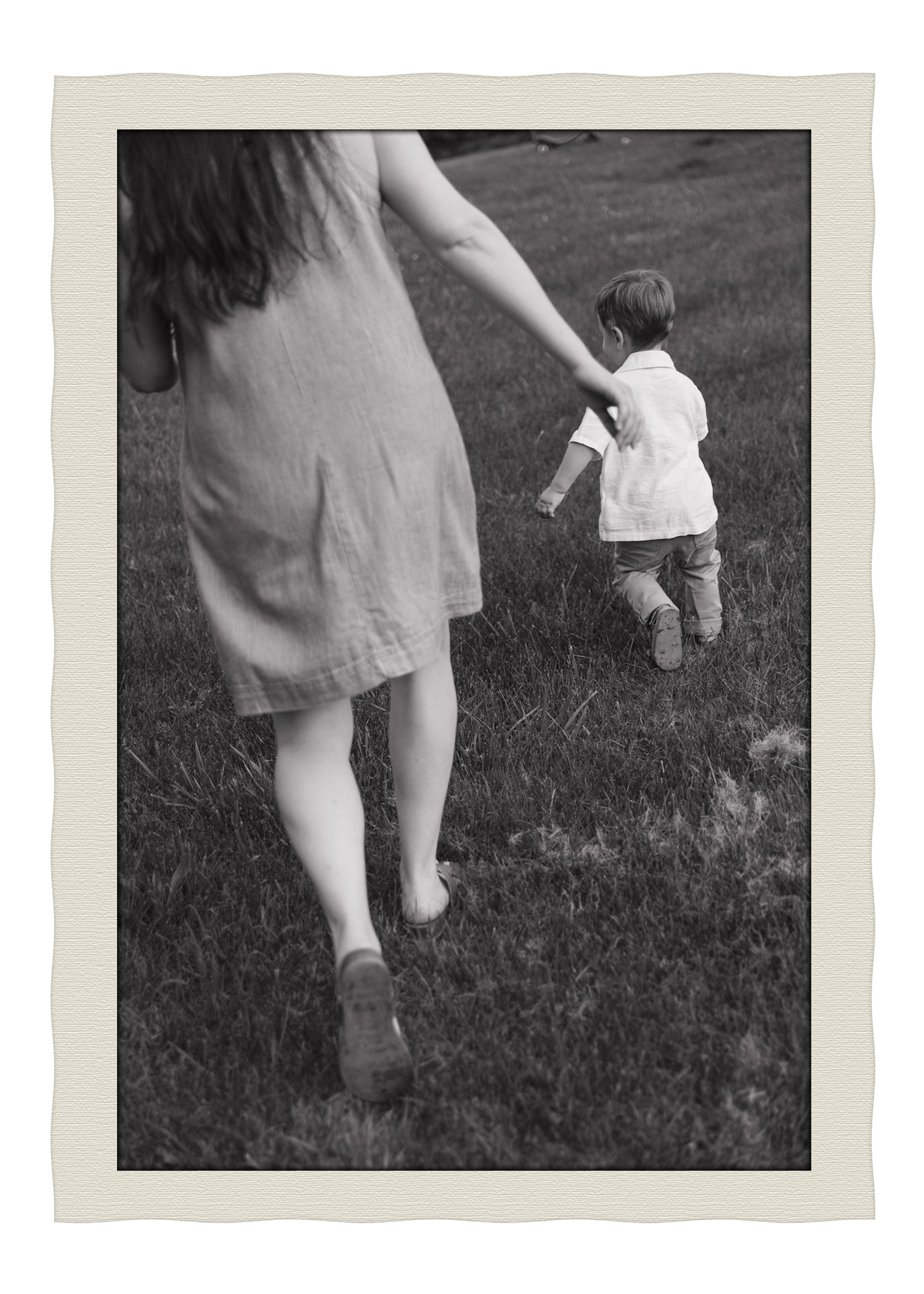 A woman and a young boy walking on grass, viewed from behind, in black and white.