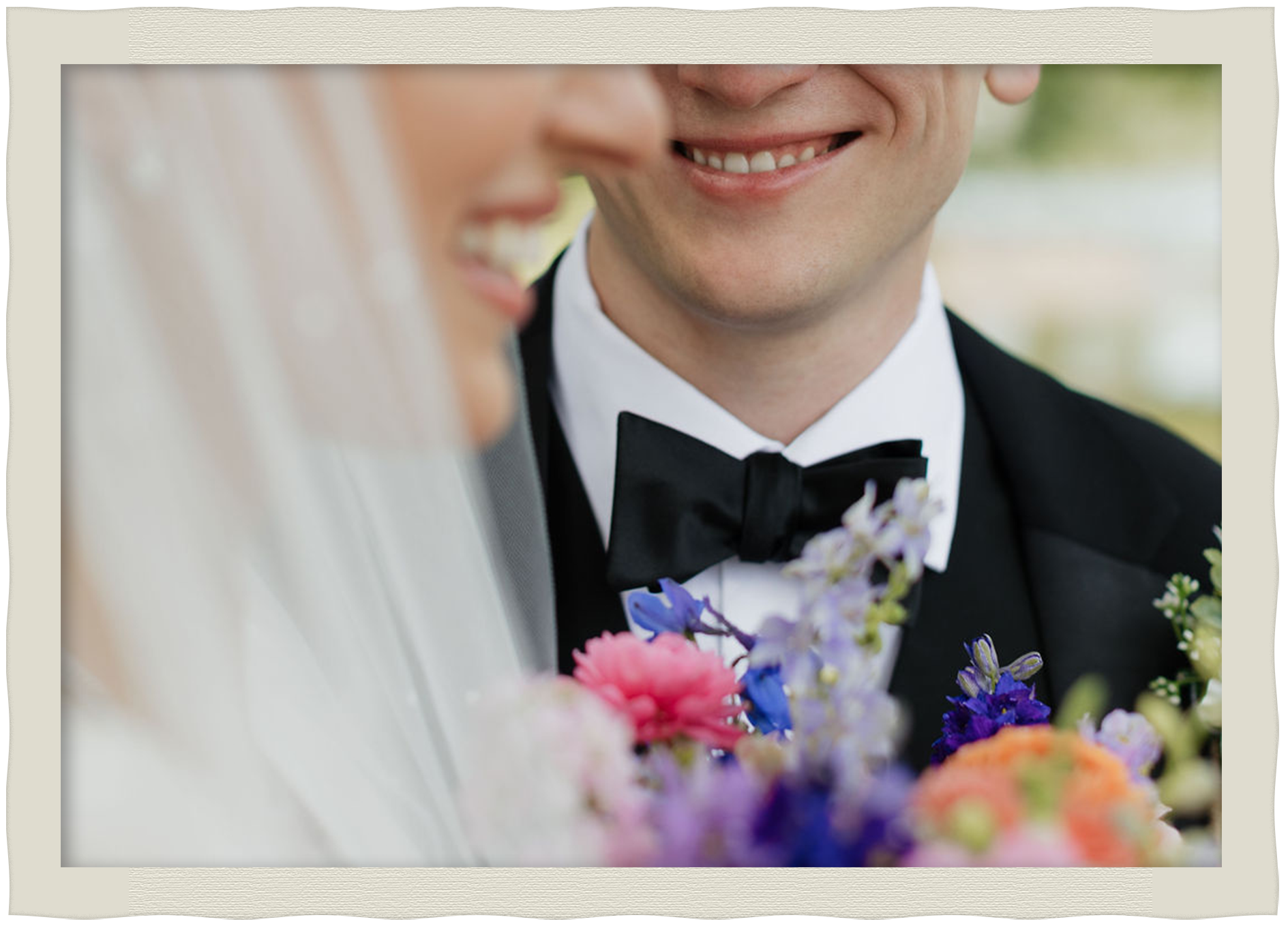 Close-up of a smiling man in a black tuxedo with a bowtie, holding a bouquet of colorful flowers, possibly a bride and groom on their wedding day.