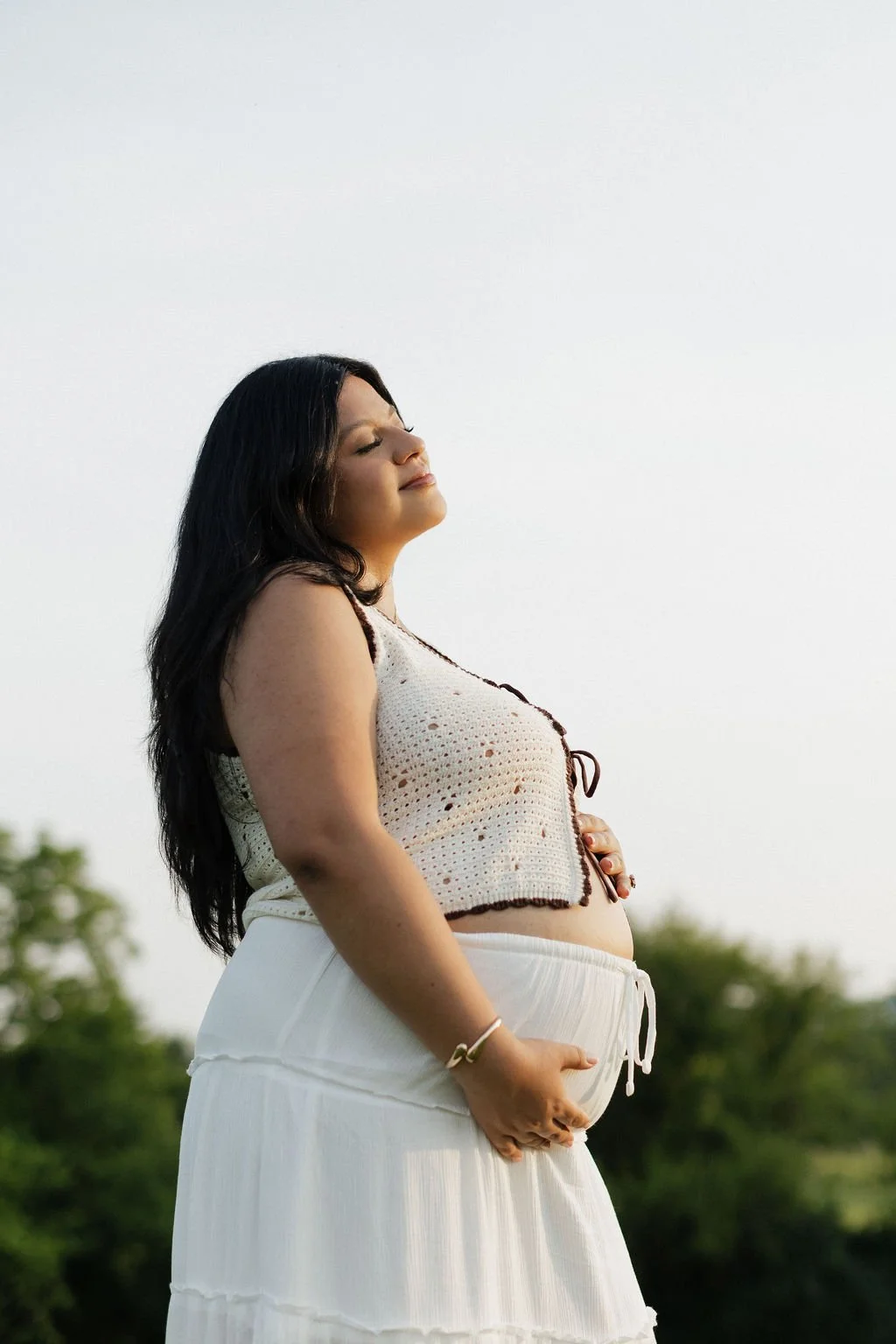A woman with long dark hair standing outdoors with eyes closed and a serene expression, holding her pregnant belly with one hand, wearing a sleeveless crochet top and white flowing skirt.