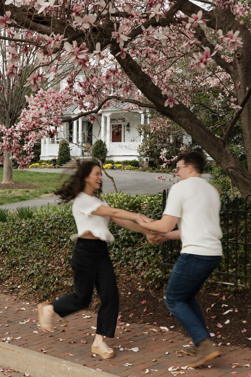 A couple dancing joyfully on a brick sidewalk under blooming cherry blossom trees with a house in the background during their engagement photo session.