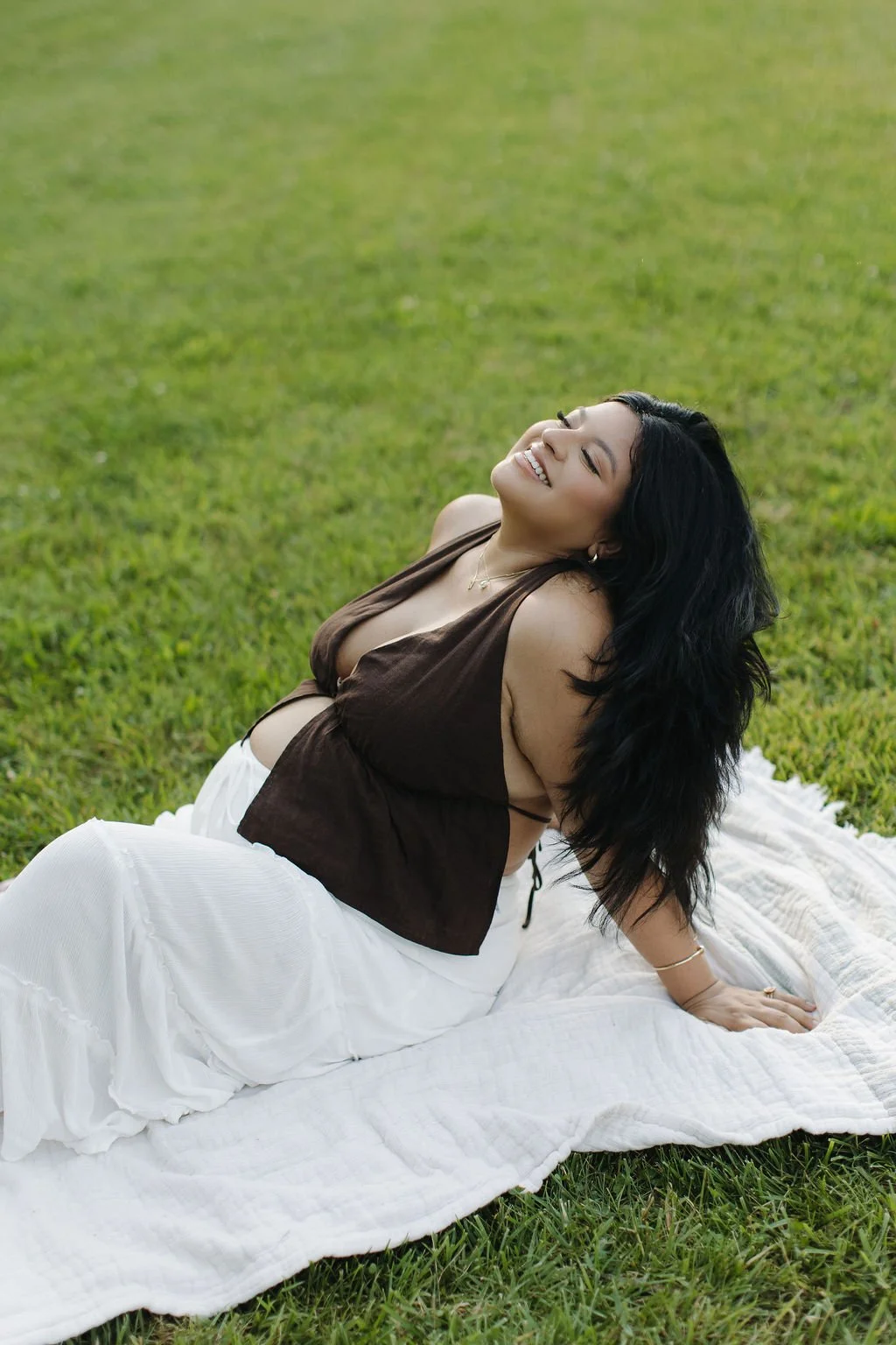 A woman with long black hair is sitting on a white blanket on green grass, smiling with her eyes closed, wearing a brown top and white skirt, enjoying a sunny day outdoors.