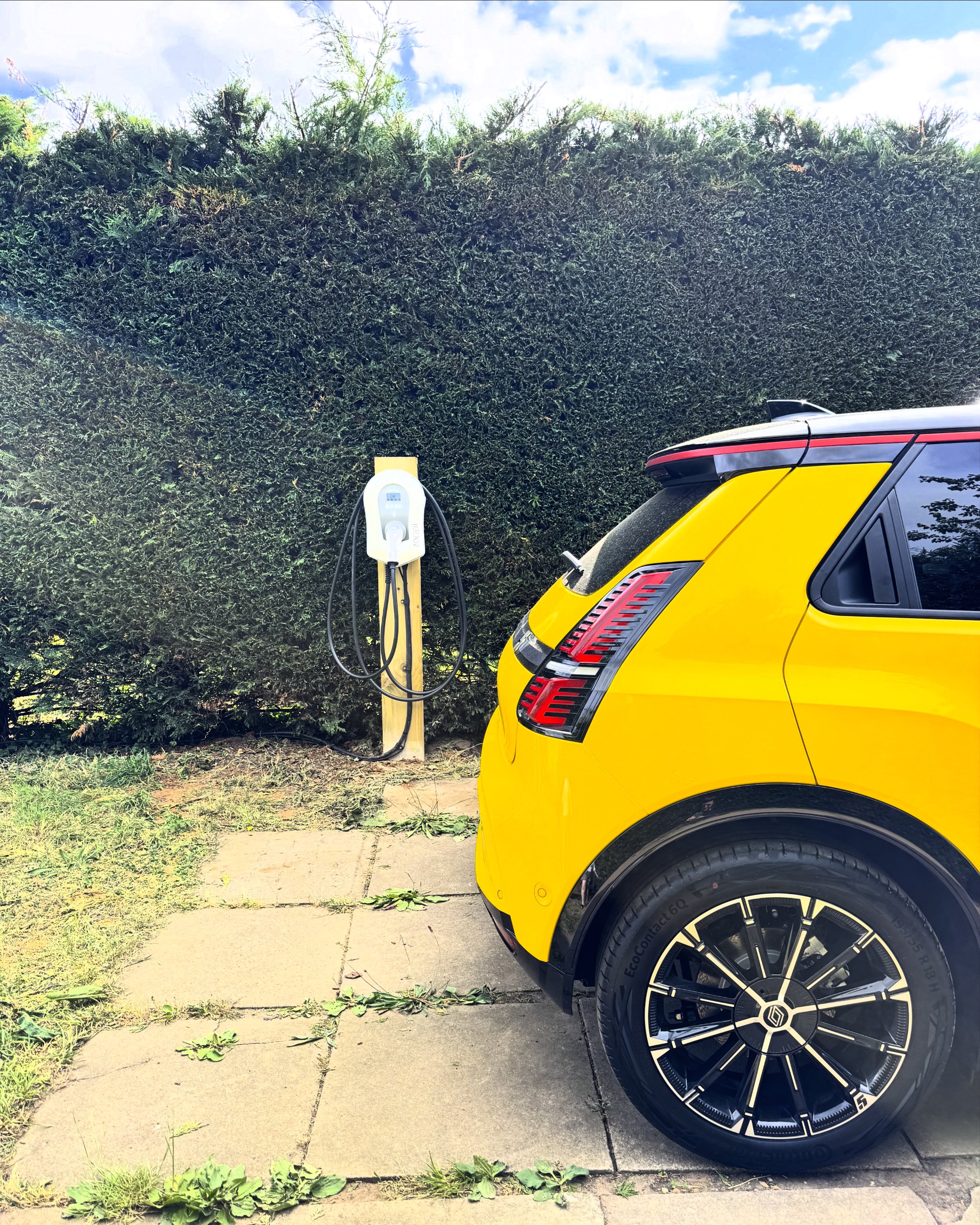 Yellow Renault 5 Electric parked on a rural driveway in front of an EV charging unit.