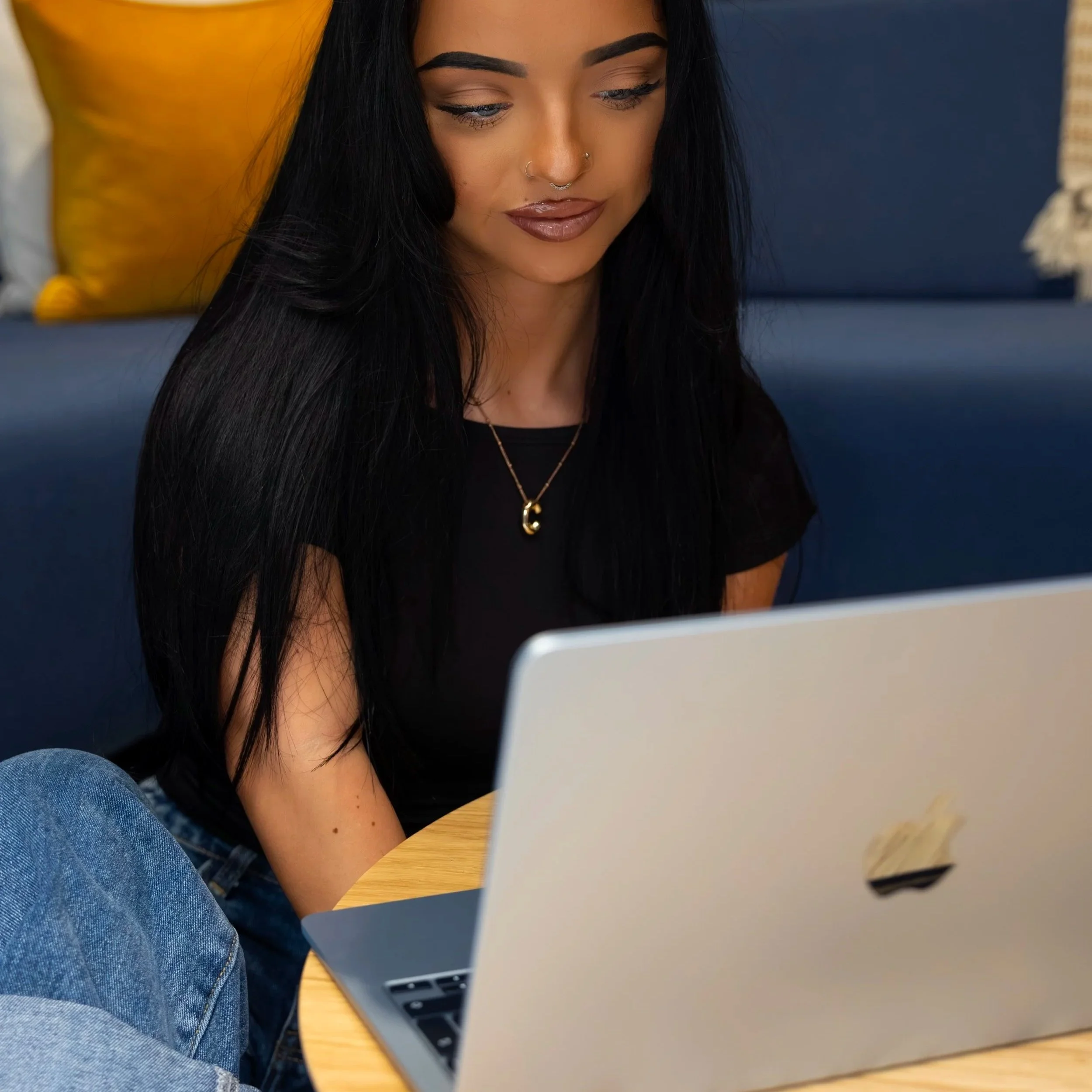A young woman with long black hair, wearing a black shirt and gold jewelry, is sitting at a round wooden table looking at a silver laptop.