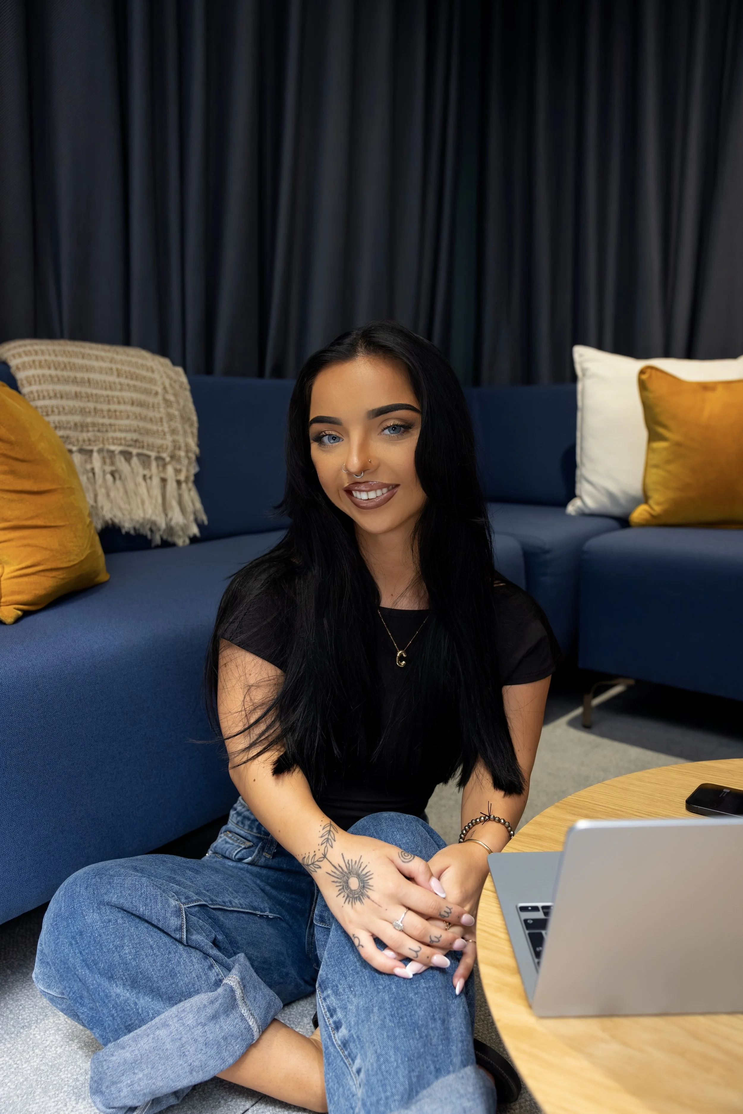 A woman with black hair and tattoos sitting cross-legged on the floor in front of a gray laptop, smiling at the camera, with a blue sofa, mustard yellow and beige cushions, gray curtains, and a wooden coffee table in the background.