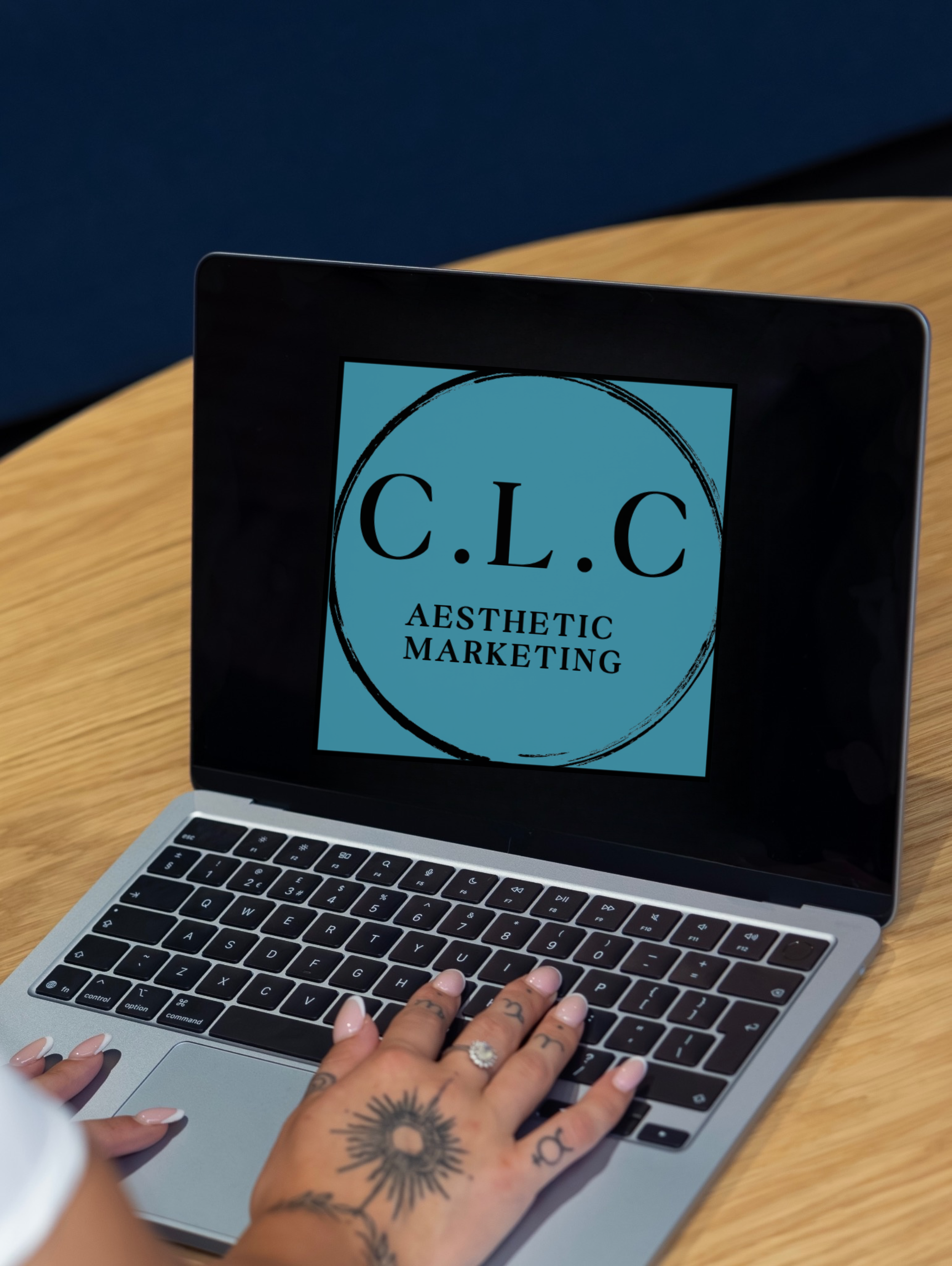 Close-up of a person with tattoos on their hand typing on a laptop with a logo reading 'C.L.C Aesthetic Marketing' on the screen.
