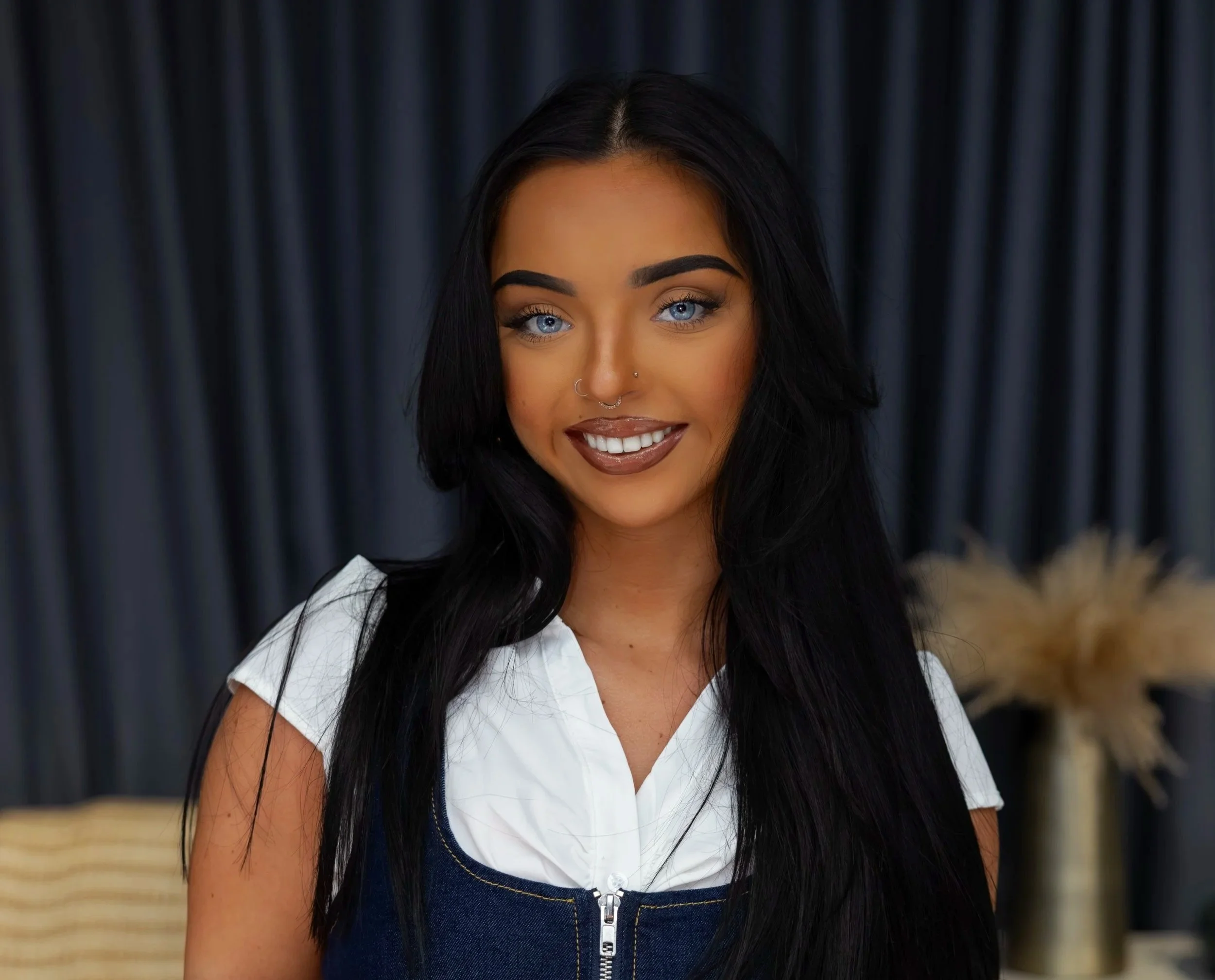 Young woman with long black hair and bright blue eyes, smiling, wearing a white top and denim overalls, with dark curtains and a vase with dried flowers in the background.