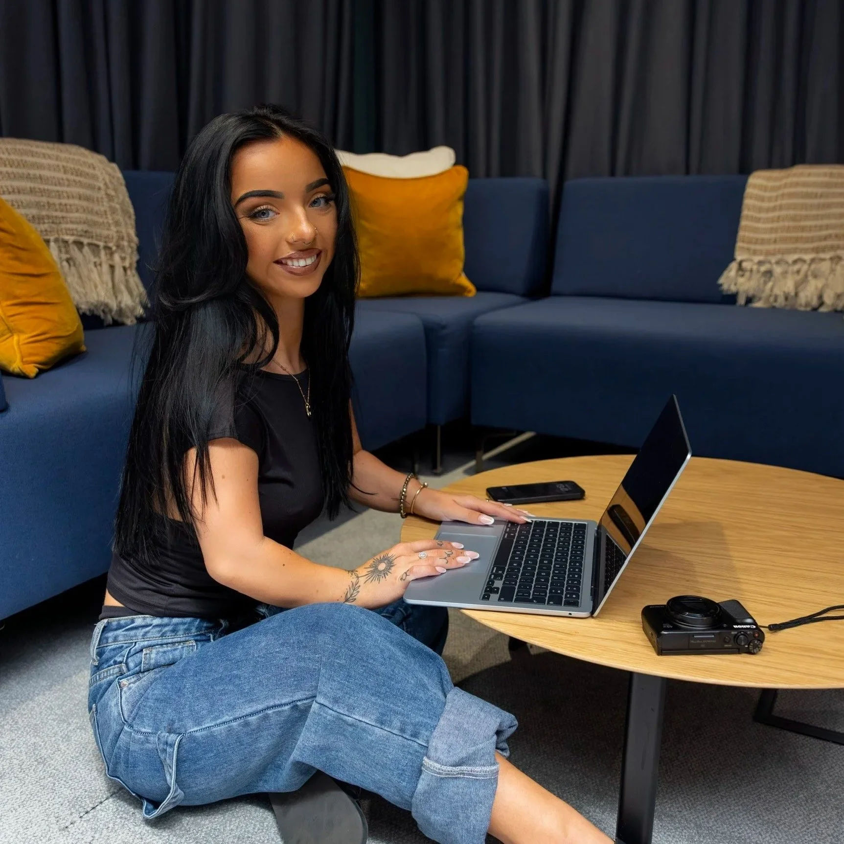 A woman with long black hair, tattoos, and wearing a black t-shirt and jeans, sitting on the floor and working on a laptop on a round wooden table, in a room with a blue sofa and mustard-colored pillows.