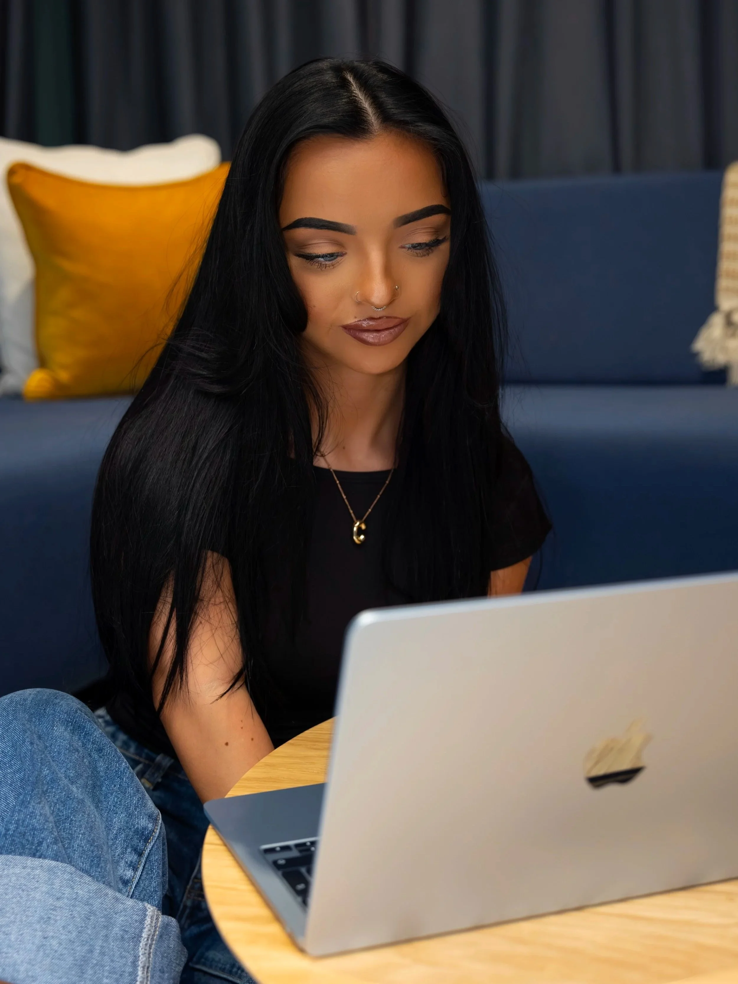 A young woman with long black hair, wearing a black shirt and a gold necklace, sitting at a round wooden table, looking at a silver laptop.