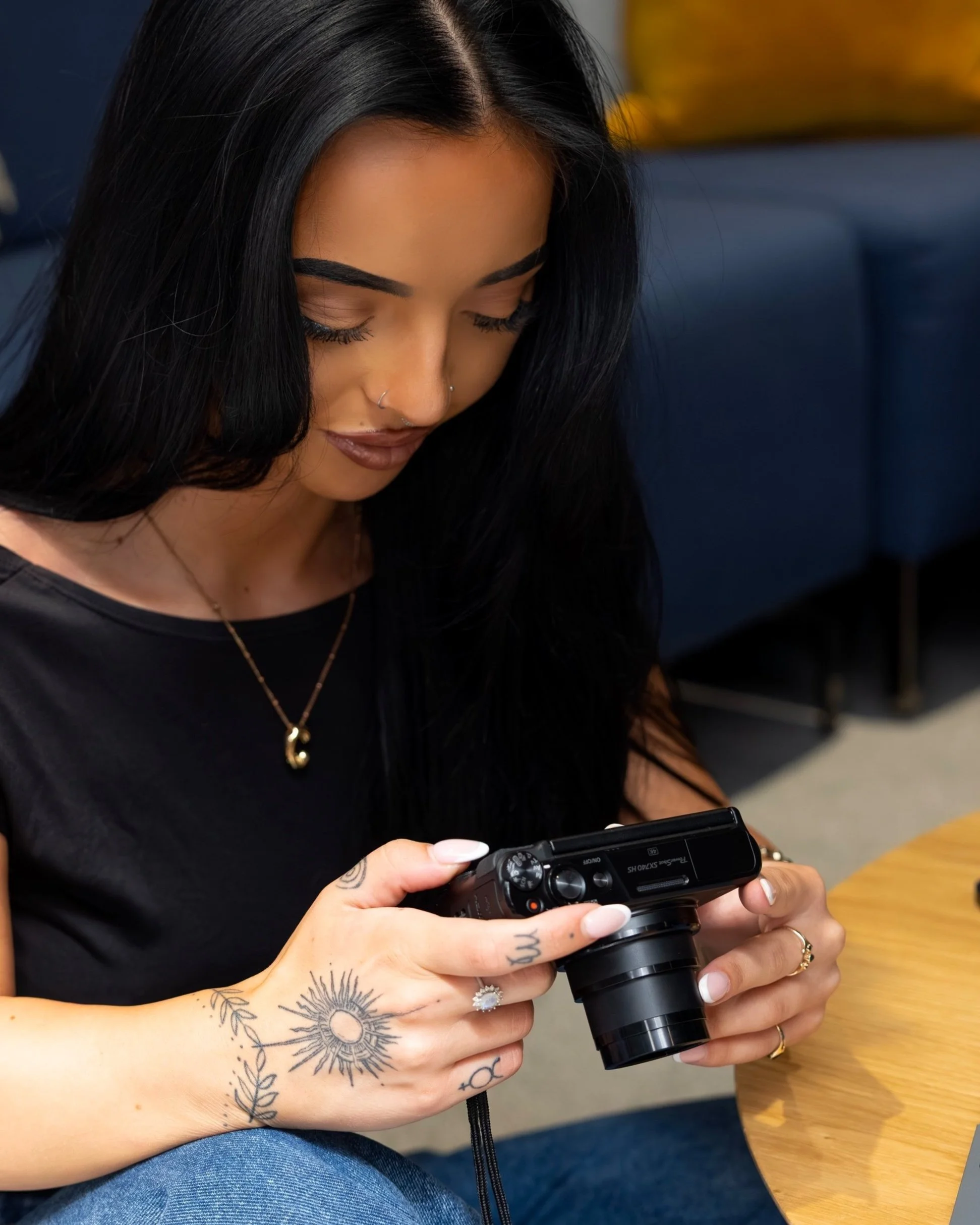 Young woman with black hair, tattoos on her hands, and jewelry, sitting at a wooden table, looking at a black digital camera in her hands.